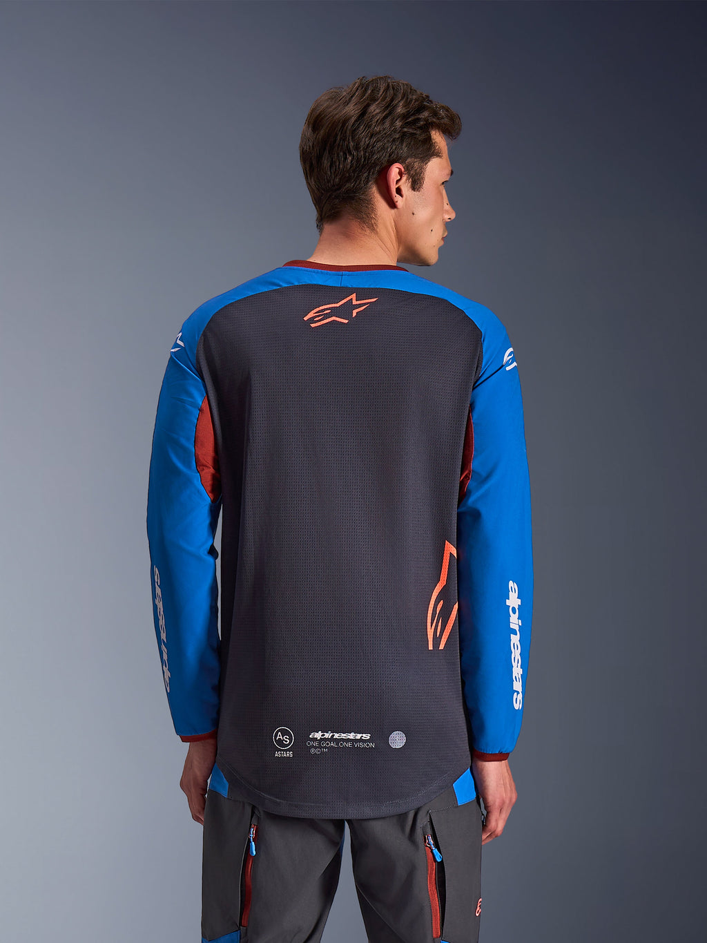 Alpinestars Maxdura Dual Jersey in donkergrijs en koningsblauw, achteraanzicht van mannelijk model, mesh torso-constructie met oranje Astars-logo, lange mouwen met witte merkdetails, bordeauxrode kraag en manchetten, gedragen met grijze technische broek, dual-sport en adventure motorkleding