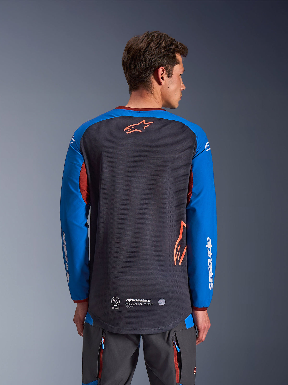 Alpinestars Maxdura Dual Jersey in donkergrijs en koningsblauw, achteraanzicht van mannelijk model, mesh torso-constructie met oranje Astars-logo, lange mouwen met witte merkdetails, bordeauxrode kraag en manchetten, gedragen met grijze technische broek, dual-sport en adventure motorkleding