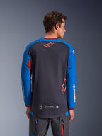 Alpinestars Maxdura Dual Jersey in donkergrijs en koningsblauw, achteraanzicht van mannelijk model, mesh torso-constructie met oranje Astars-logo, lange mouwen met witte merkdetails, bordeauxrode kraag en manchetten, gedragen met grijze technische broek, dual-sport en adventure motorkleding