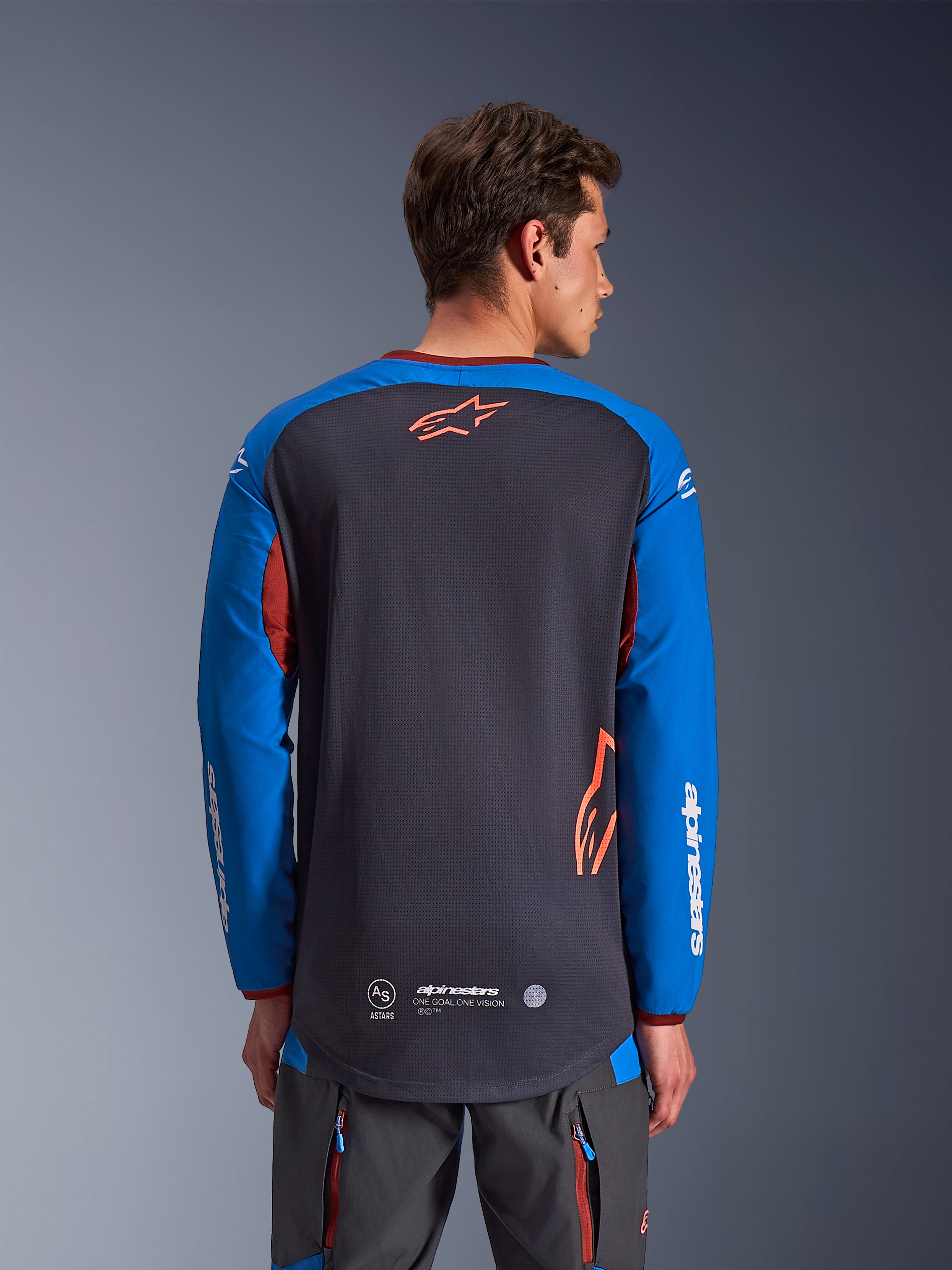 Alpinestars Maxdura Dual Jersey in donkergrijs en koningsblauw, achteraanzicht van mannelijk model, mesh torso-constructie met oranje Astars-logo, lange mouwen met witte merkdetails, bordeauxrode kraag en manchetten, gedragen met grijze technische broek, dual-sport en adventure motorkleding