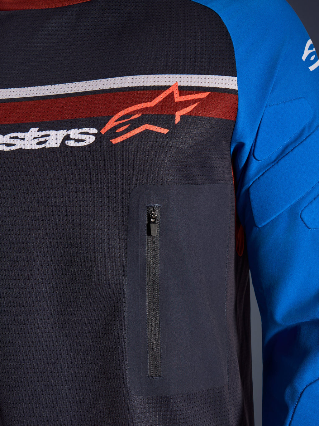 Alpinestars Maxdura Dual Jersey, detailopname van borst en mouw, donkergrijs en koningsblauw, voorzien van een borstzak met ritssluiting, ademende mesh torso en geïntegreerde padding op de mouwen, met het Alpinestars-logo over rode en witte strepen.
