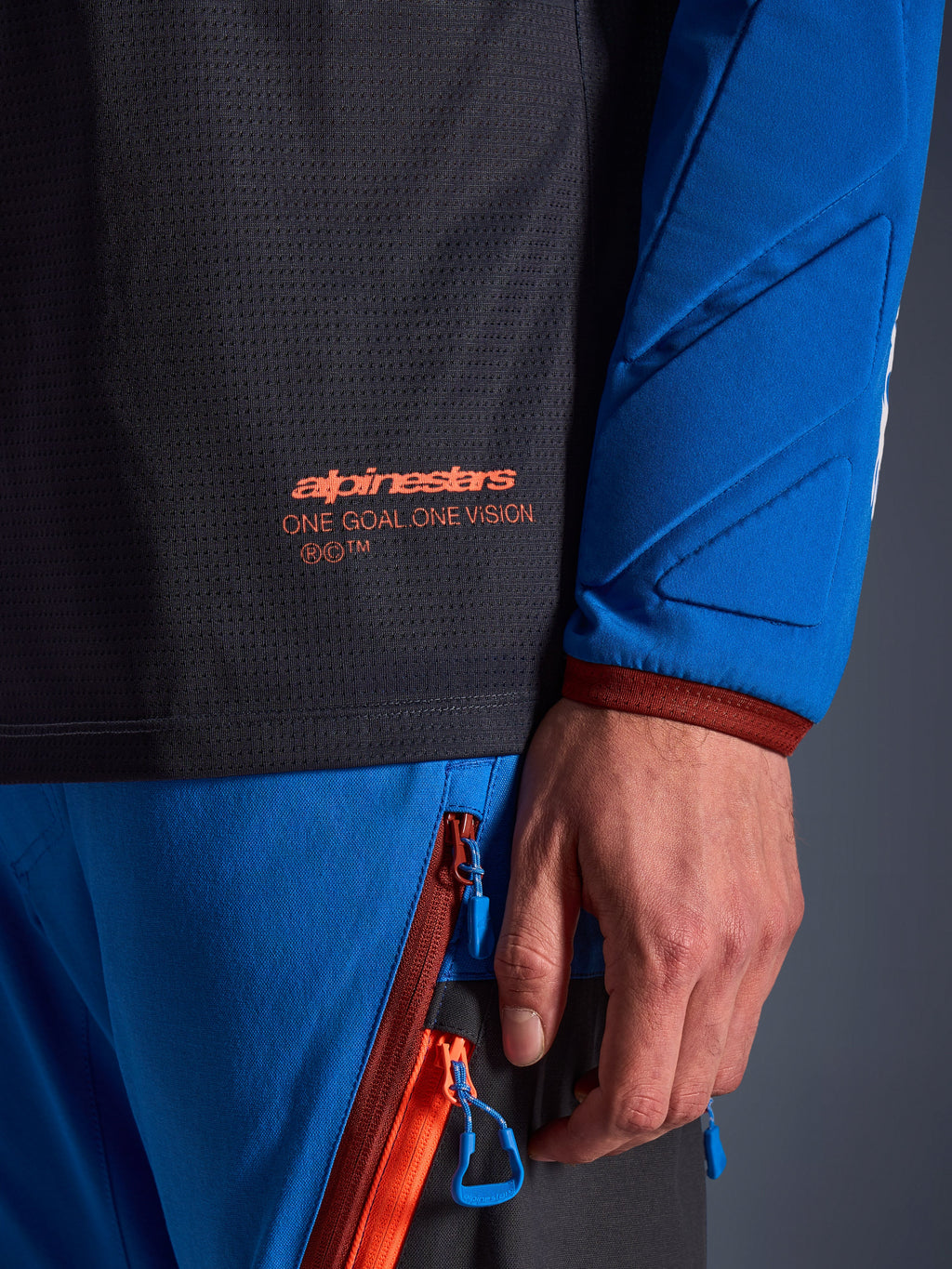 Close-up van de Alpinestars Maxdura Dual Jersey in Deep Gray en Royal Blue, met een oranje logo en de tekst 'One Goal One Vision' op de mesh romp, gewatteerde mouw met verstevigde geometrische panelen, technisch detail van adventure-rijuitrusting