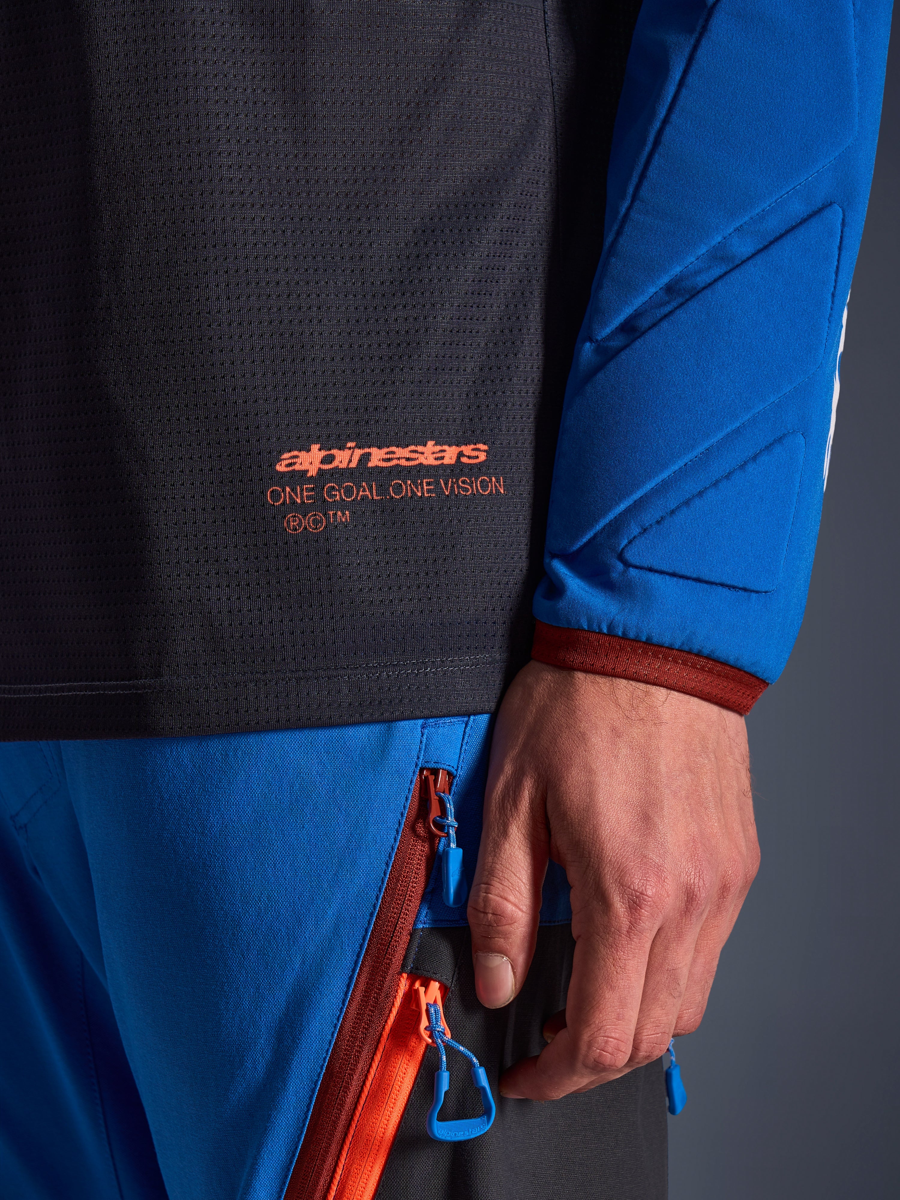 Close-up van de Alpinestars Maxdura Dual Jersey in Deep Gray en Royal Blue, met een oranje logo en de tekst 'One Goal One Vision' op de mesh romp, gewatteerde mouw met verstevigde geometrische panelen, technisch detail van adventure-rijuitrusting