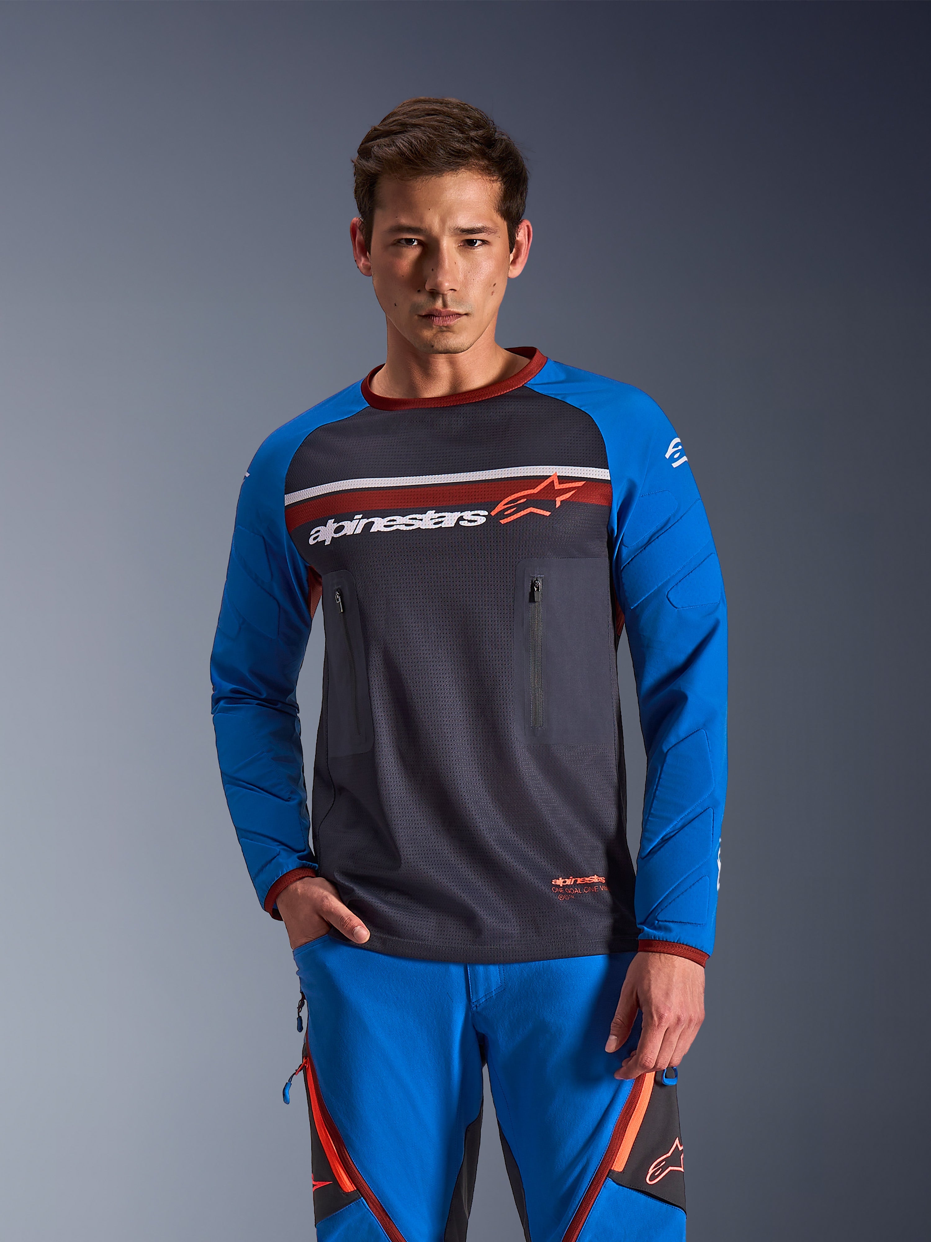 Alpinestars Maxdura Dual Jersey, motor jersey met lange mouwen, donkergrijs en koningsblauw met rode accenten, voorzien van borstzakken met rits en gevoerde mouwen, gedragen met bijpassende blauwe broek, technische kleding voor adventure en dual-sport rijden
