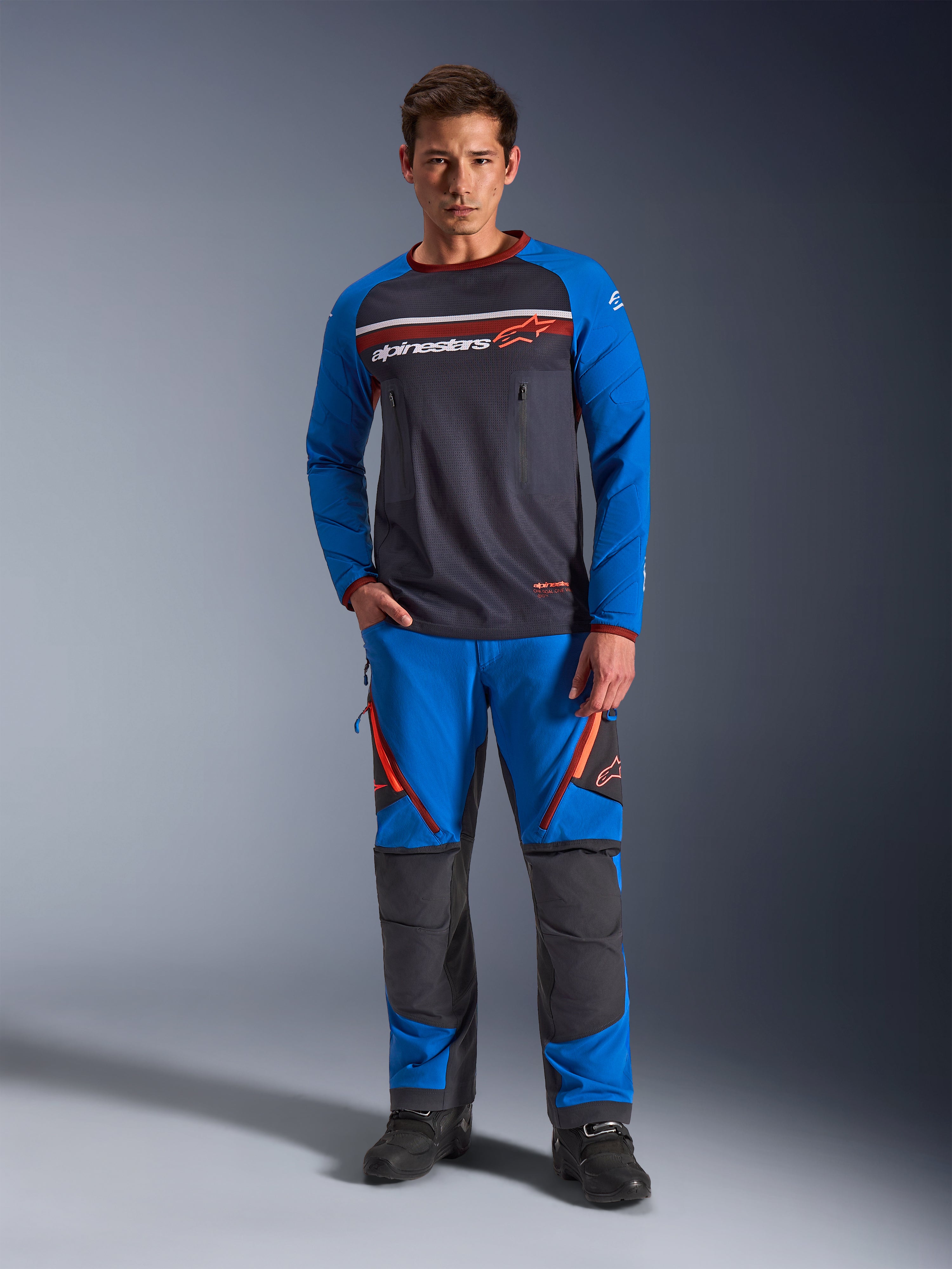 Alpinestars Maxdura Dual Jersey, motor jersey, donkergrijs en koningsblauw, volledige mesh torso met gewatteerde mouwen en borstzakken met rits, voorzien van wit en oranje Alpinestars-logo, gedragen met bijpassende adventure broek en zwarte laarzen