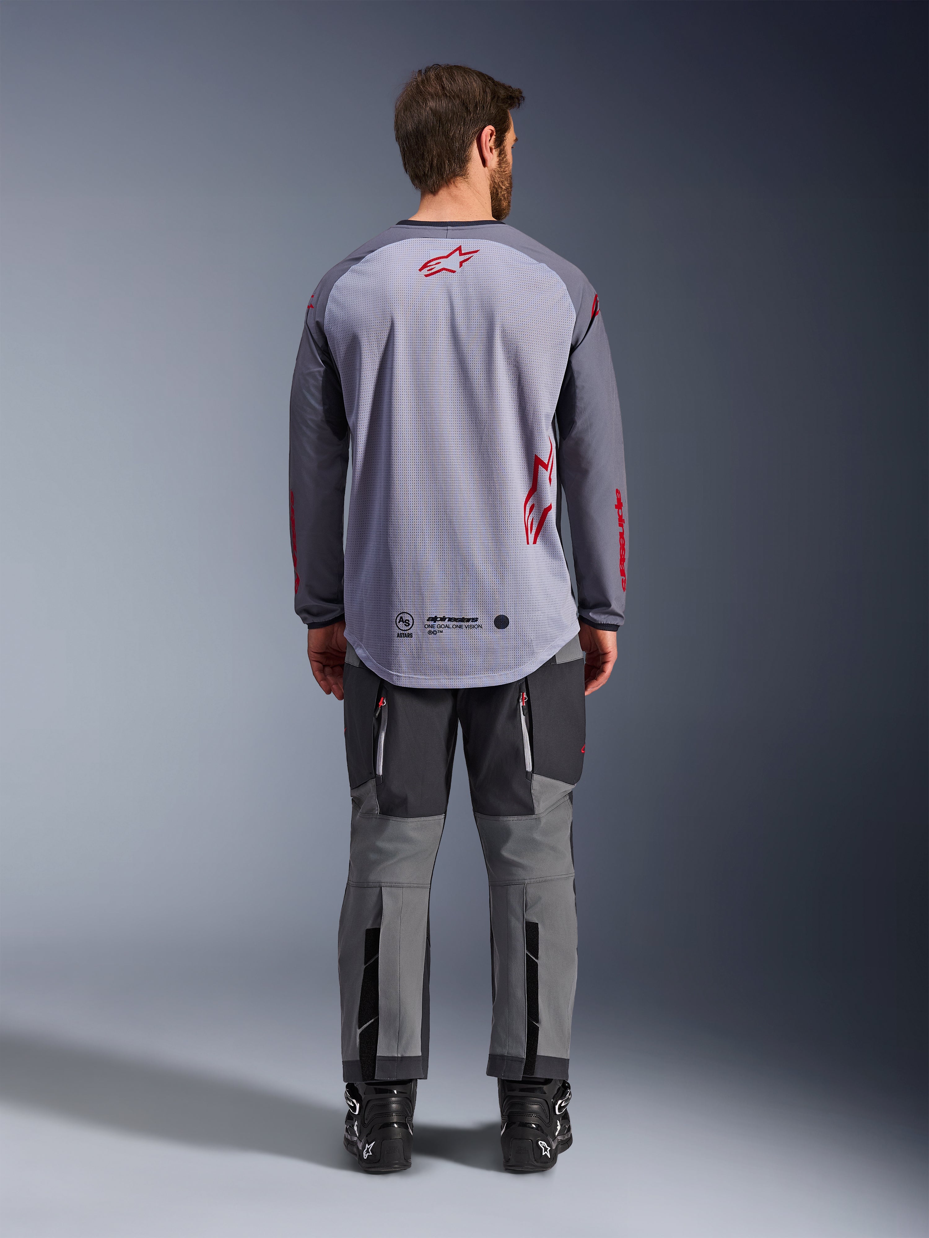 Mannelijk model van achteren gezien die de Alpinestars Maxdura Dual Jersey draagt, grijs en donkergrijs adventure-shirt met rode logo-accenten, mesh rugconstructie, gedragen met bijpassende grijze off-road broek en zwarte laarzen.