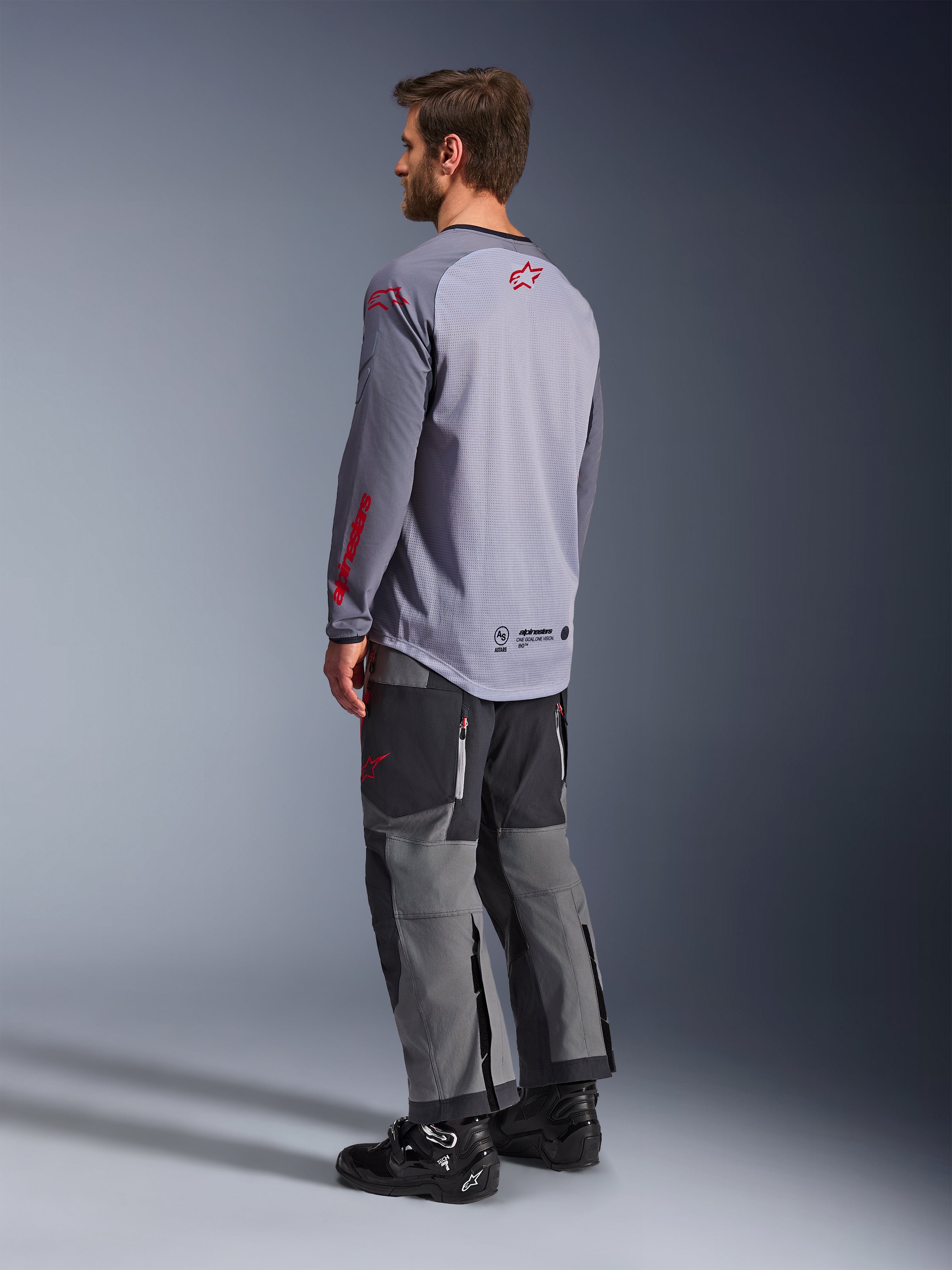 Man die een Alpinestars Maxdura Dual Jersey draagt, lichtgrijs met rode Alpinestars-logo's, achteraanzicht met mesh torso en lange mouwen, gecombineerd met een antracietgrijze technische broek en zwarte motorlaarzen