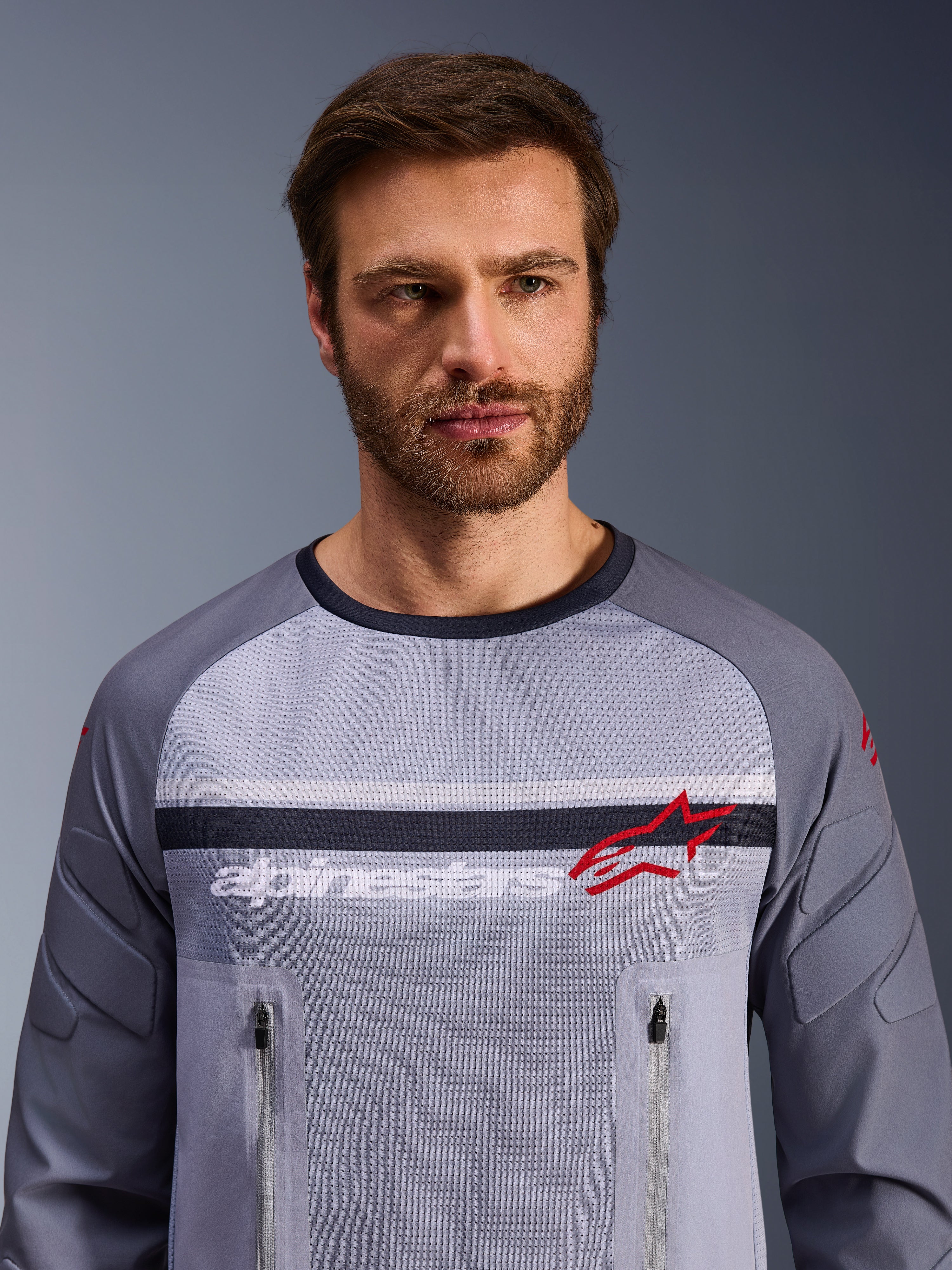 Alpinestars Maxdura Dual Jersey, midden-grijs en donkergrijs, gedragen door mannelijk model, voorzien van een mesh torso voor ventilatie, dubbele borstzakken met ritssluiting, witte 'alpinestars' branding en rood ster-logo, hoogwaardige off-road adventure uitrusting