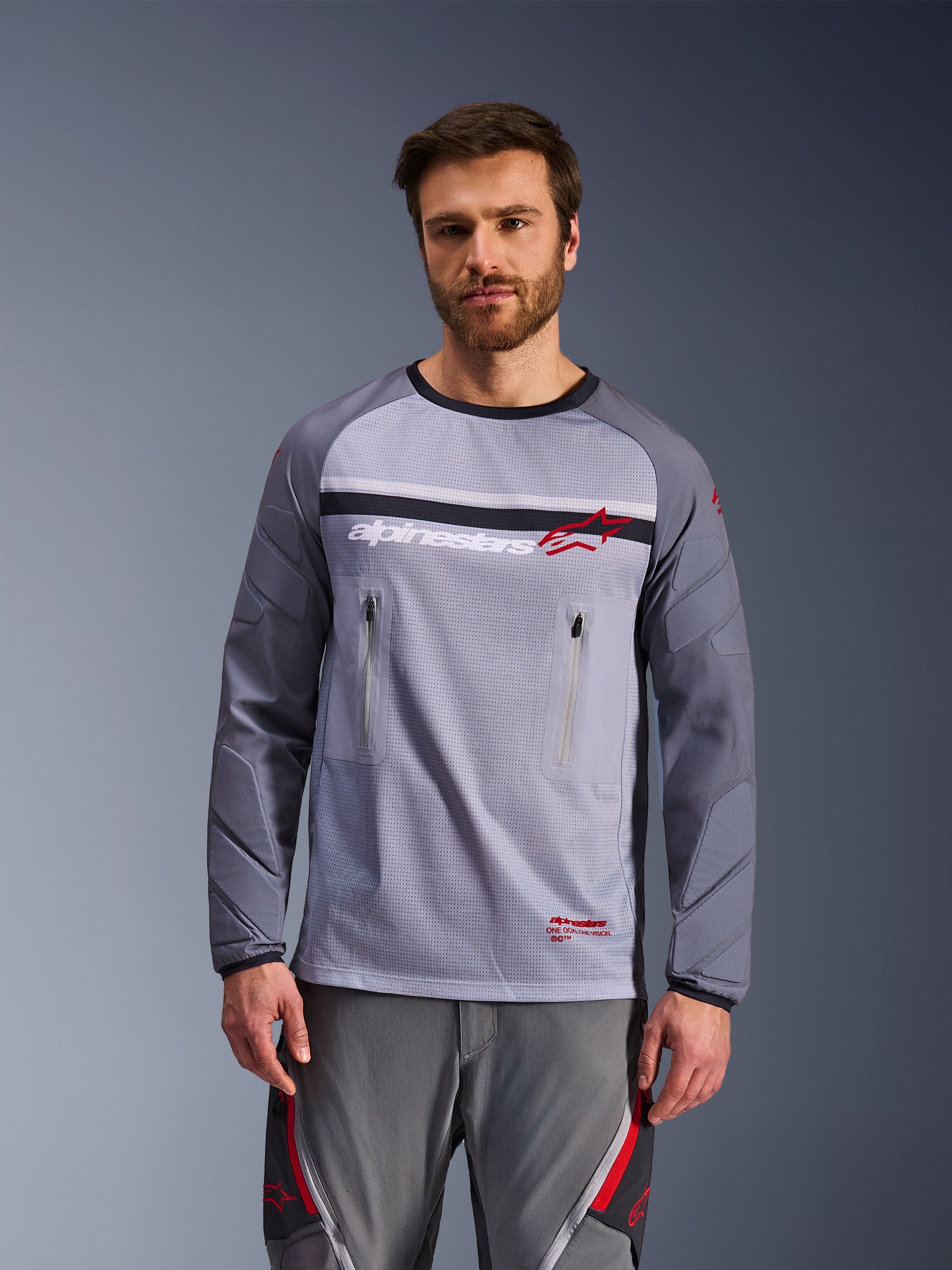 Alpinestars Maxdura Dual Jersey, midden- en donkergrijs, gedragen door een model met bijpassende grijze broek, voorzien van borstzakken met rits, mesh torso-constructie en gevoerde slijtvaste mouwen, adventure motor off-road uitrusting