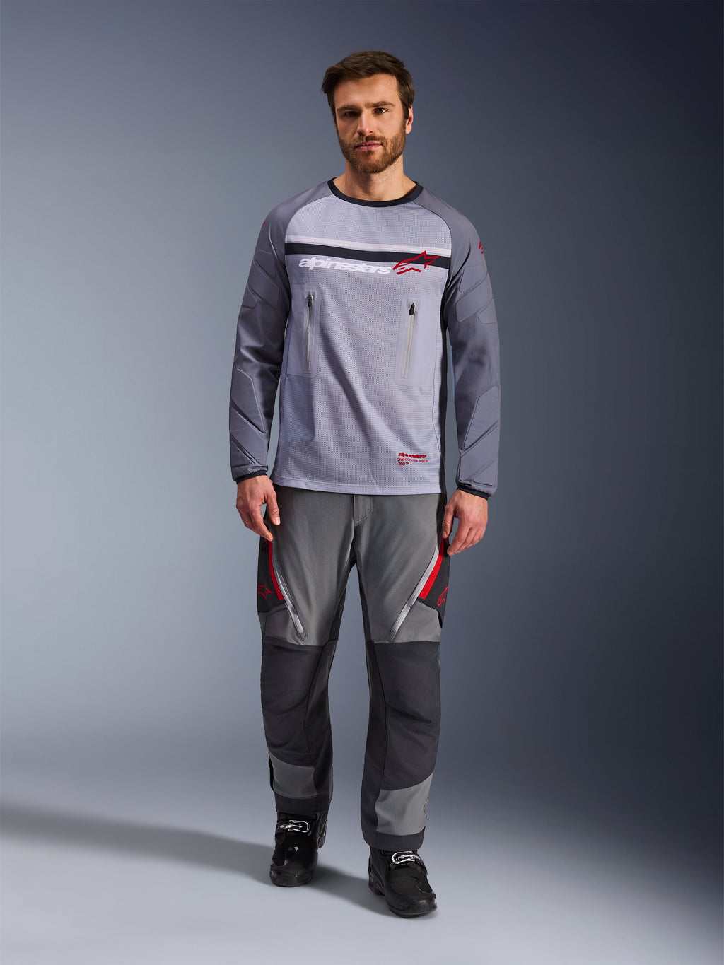 Alpinestars Maxdura Dual Jersey, Mid Gray en Dark Gray, adventure motor jersey met mesh torso en borstzakken met rits, slijtvaste mouwen met geïntegreerde padding, gedragen met bijpassende grijze performance broek met rode accenten, technische off-road motorkleding