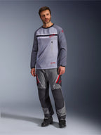 Een man die de Alpinestars Maxdura Dual Jersey in Mid Gray en Dark Gray draagt, gecombineerd met een bijpassende grijze en zwarte adventure-broek en zwarte laarzen, voorzien van een ademende mesh torso, borstzakken met ritssluiting en rode logo-accenten.