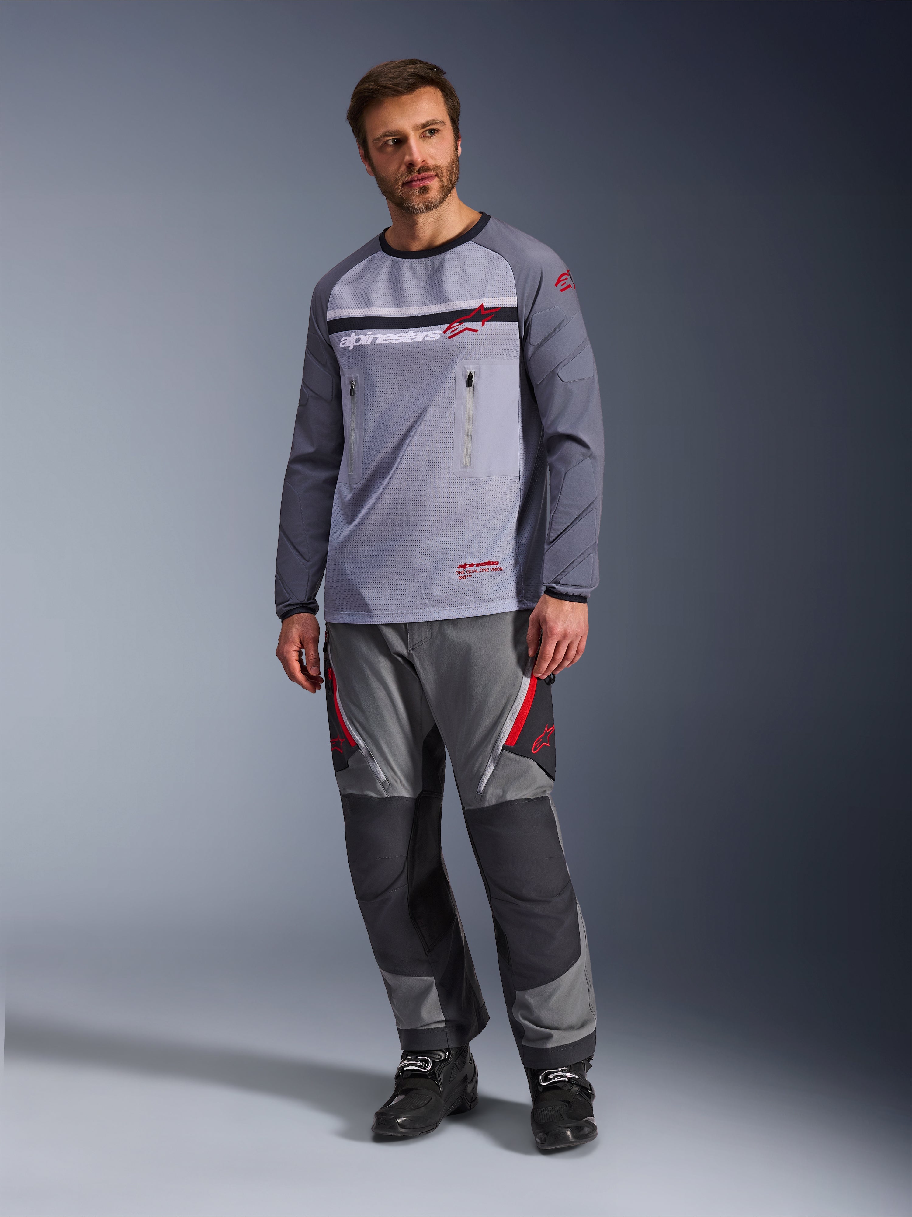 Een man die de Alpinestars Maxdura Dual Jersey in Mid Gray en Dark Gray draagt, gecombineerd met een bijpassende grijze en zwarte adventure-broek en zwarte laarzen, voorzien van een ademende mesh torso, borstzakken met ritssluiting en rode logo-accenten.