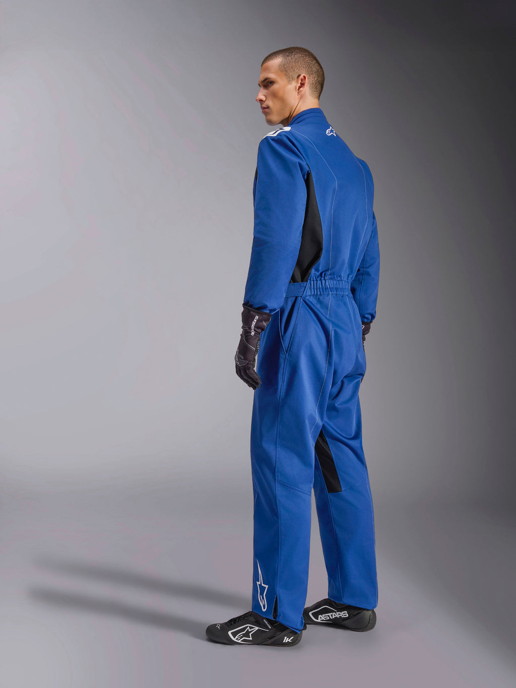 Alpinestars Kart Indoor V2 Suit, blauw kartpak, achter- en zijaanzicht, eendelig racepak met zwarte zijpanelen en accenten aan de binnenzijde van de benen, elastische taille en witte logo's, gedragen met zwarte racehandschoenen en schoeisel
