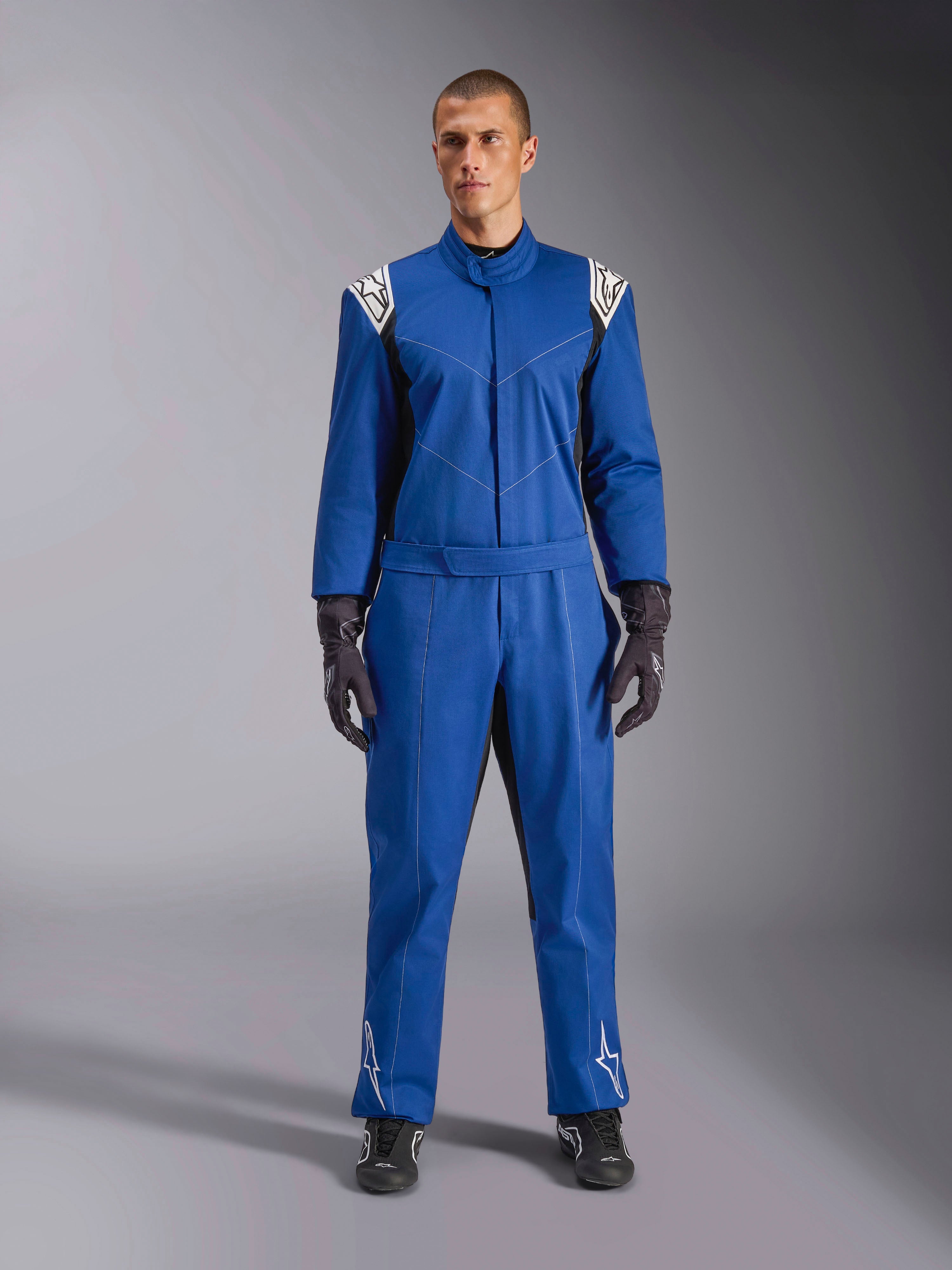 Mannelijk model in een blauw Alpinestars Kart Indoor V2-pak, eendelig kartpak met witte sterlogo's op schouders en schenen, witte contraststiksels en een elastische tailleriem, gestyled met zwarte racehandschoenen en schoenen, ontworpen voor recreatief circuitgebruik