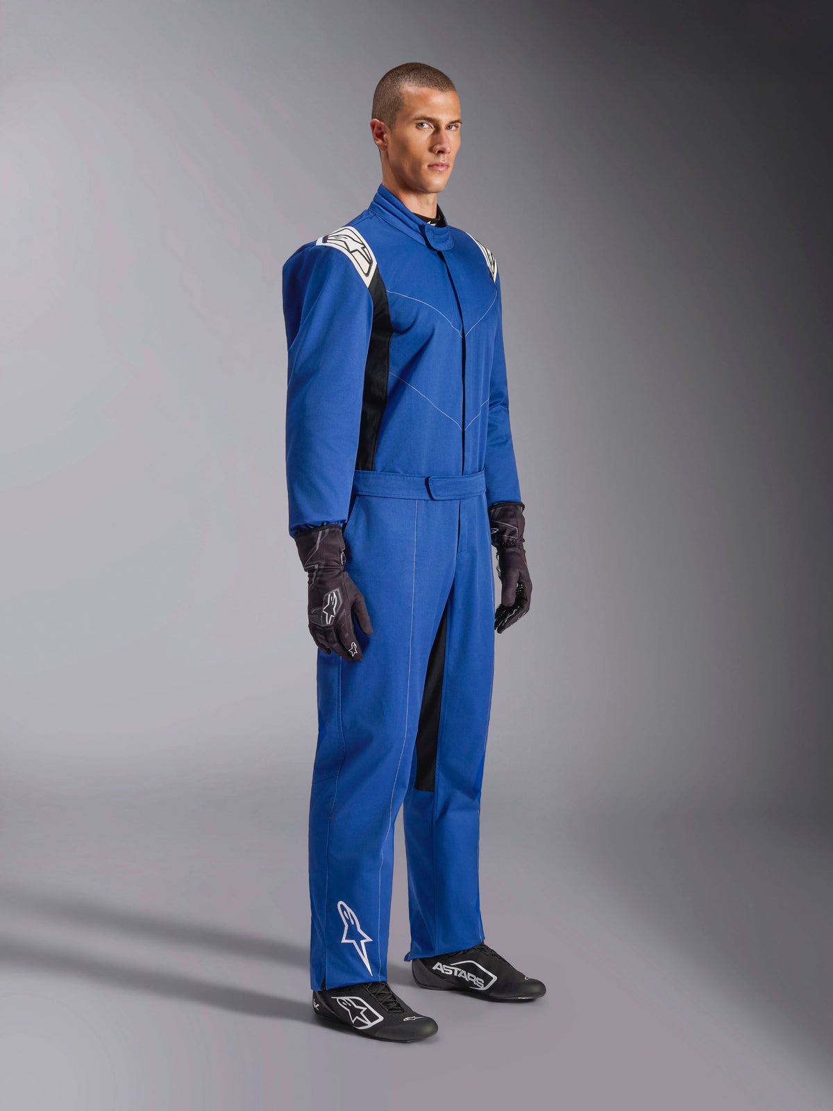 Alpinestars Kart Indoor V2 Suit, blauw kartpak, eendelige race-overall met witte schouderpanelen en logo's, verstelbare tailleband, wit logo op het onderbeen, gedragen door model met zwarte racehandschoenen en schoenen, motorsportkleding voor indoor karten