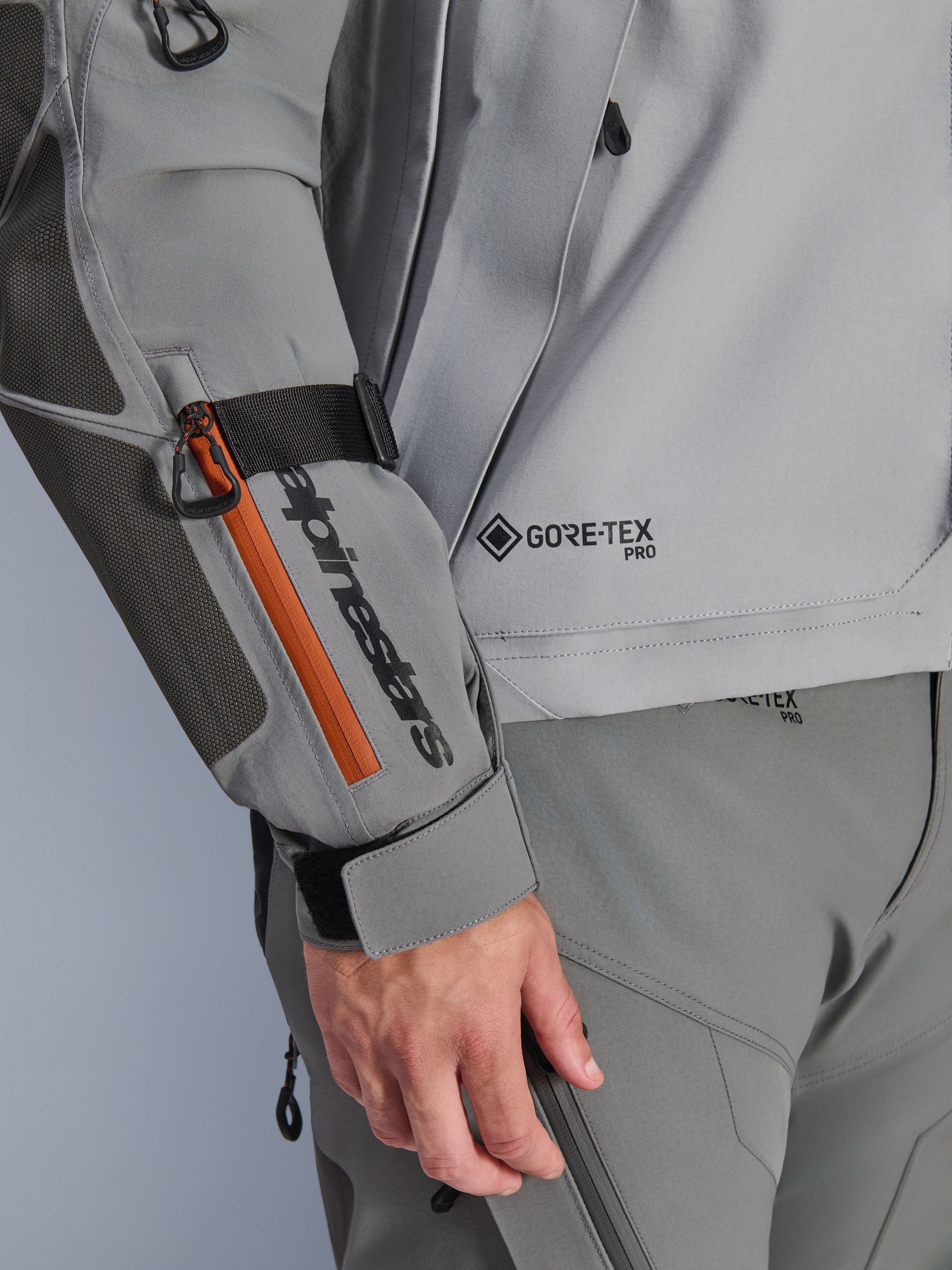 Alpinestars Nazca 3L GORE-TEX PRO Jas, donkergrijze en middengrijze adventure jas, close-up detail van mouw met zwarte merkdetails en oranje ritsaccent, GORE-TEX PRO-logo op de torso, duurzame textielconstructie met versterkt elleboogstuk