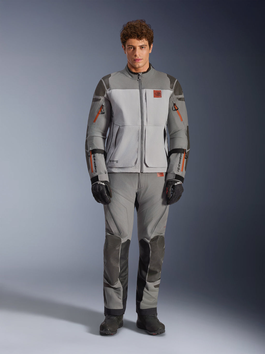 Alpinestars Nazca 3L GORE-TEX PRO Jas, adventure motorjas, donkergrijs en middengrijs, gedragen door een model met bijpassende broek en zwarte handschoenen, voorzien van oranje ritsaccenten en rood logo op de borst, technische beschermende uitrusting voor extreem rijden