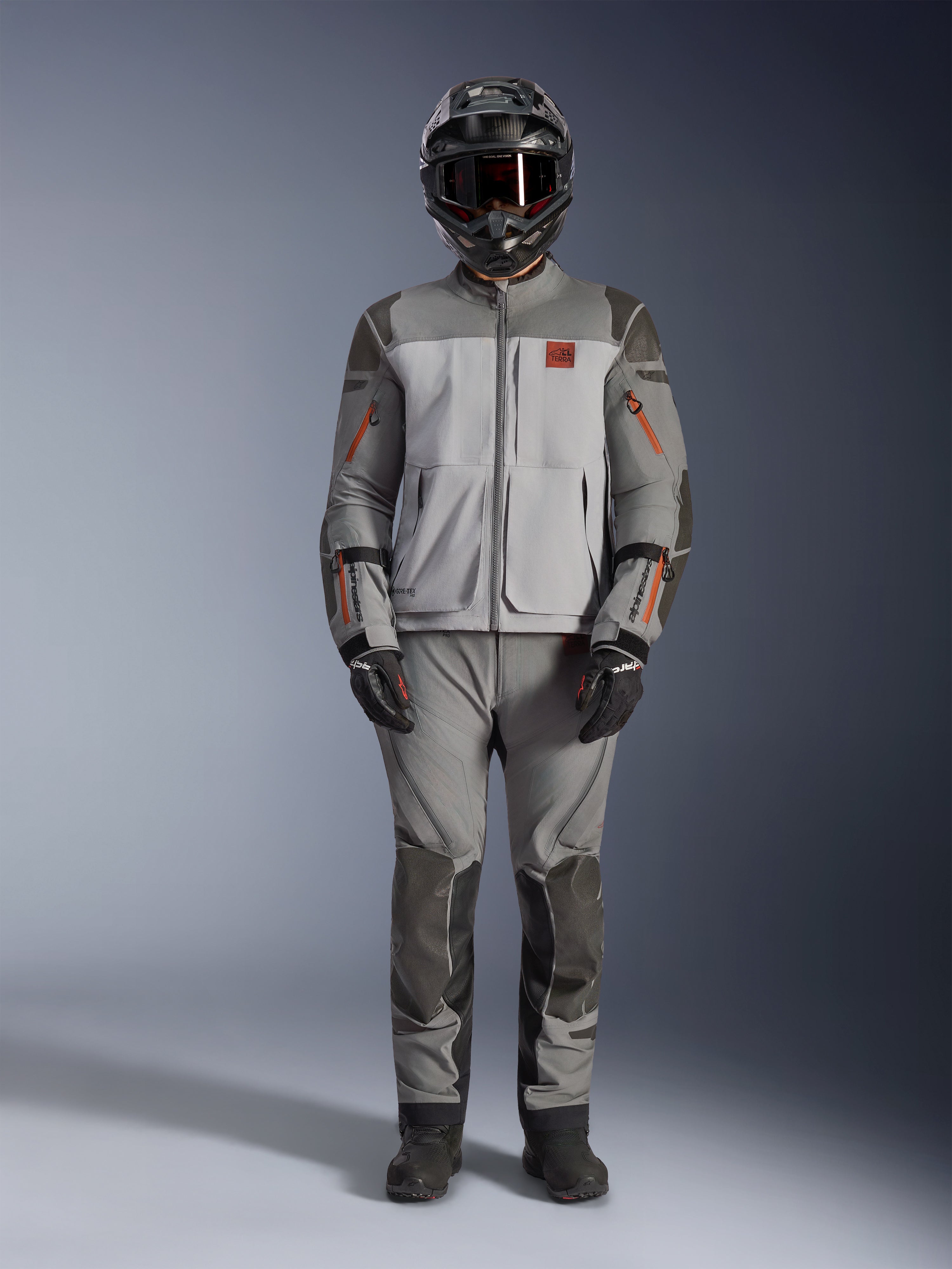 Alpinestars Nazca 3L GORE-TEX PRO Jas, adventure motorjas, donkergrijs en middengrijs met oranje accenten, voorzien van Superfabric-versterkingen en ventilatieritsen aan de voorzijde, gedragen met bijpassende broek en zwarte helm, technische all-terrain motoruitrusting