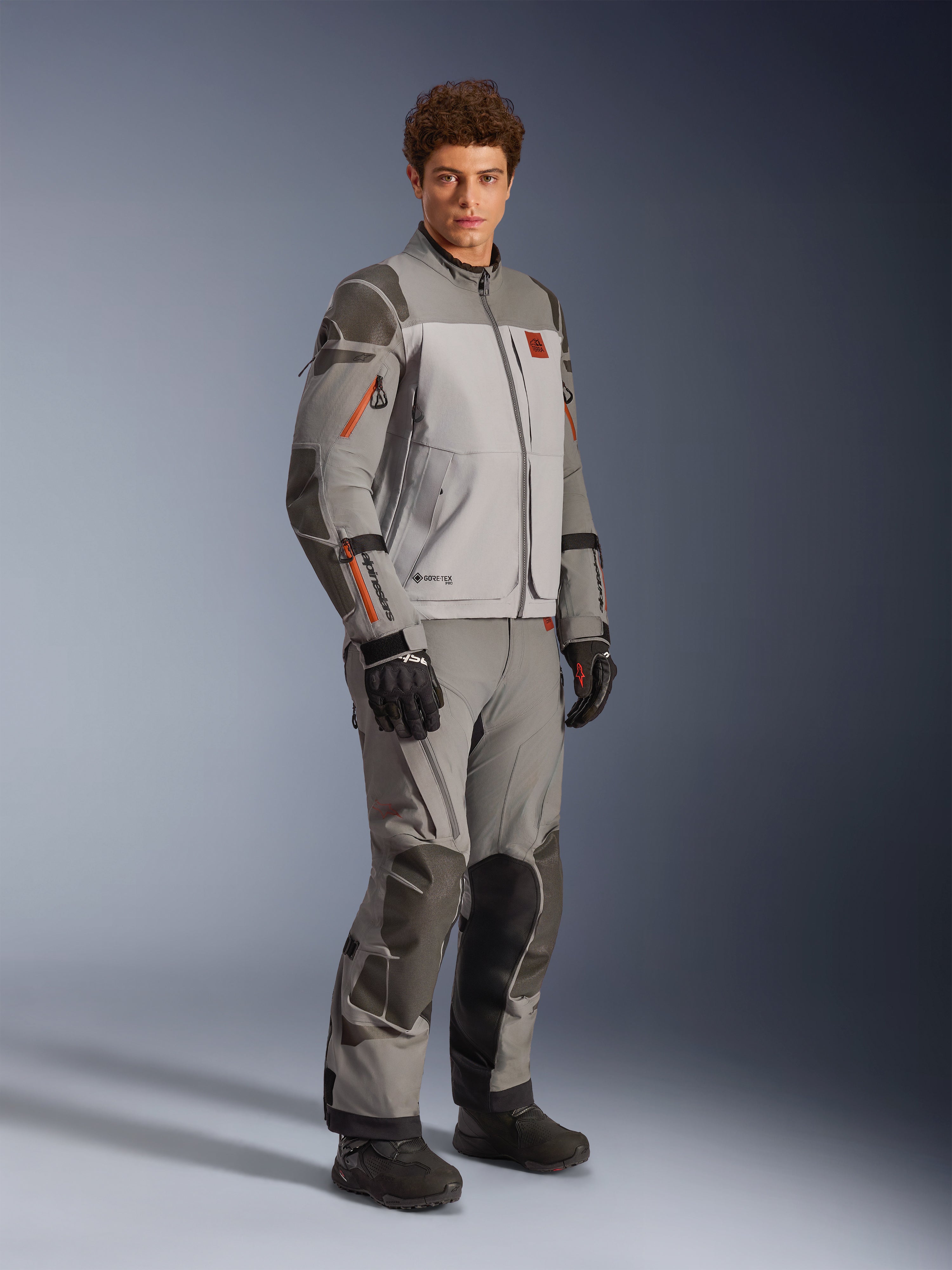Alpinestars Nazca 3L GORE-TEX PRO Jas in Dark Gray Mid Gray, adventure motorrij-outfit gedragen met bijpassende broek en zwarte handschoenen, voorzien van oranje ritsaccenten, beschermende Superfabric®-versterkingen en GORE-TEX-branding.