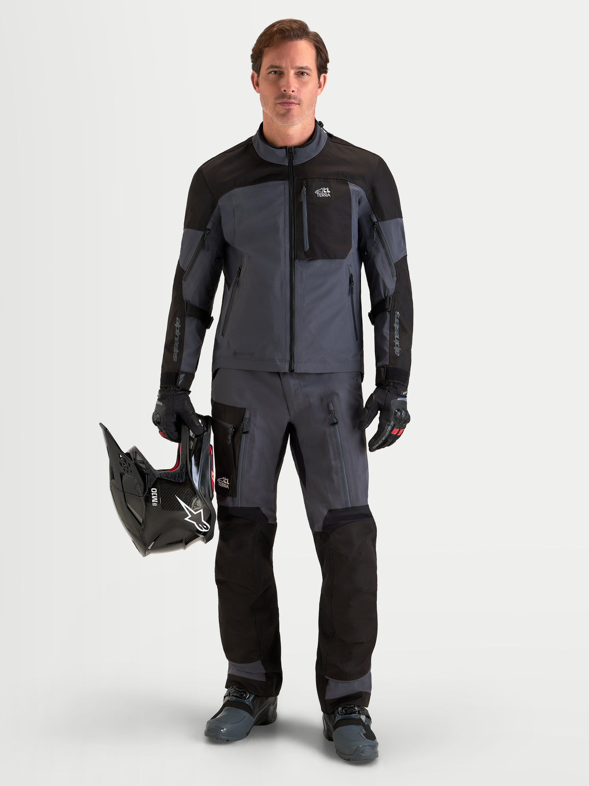 Alpinestars Arenis Gore-Tex jas, adventure motorjas voor heren in zwart en donkergrijs, gedragen door een rijder die een helm vasthoudt, met waterdichte GORE-TEX textielconstructie