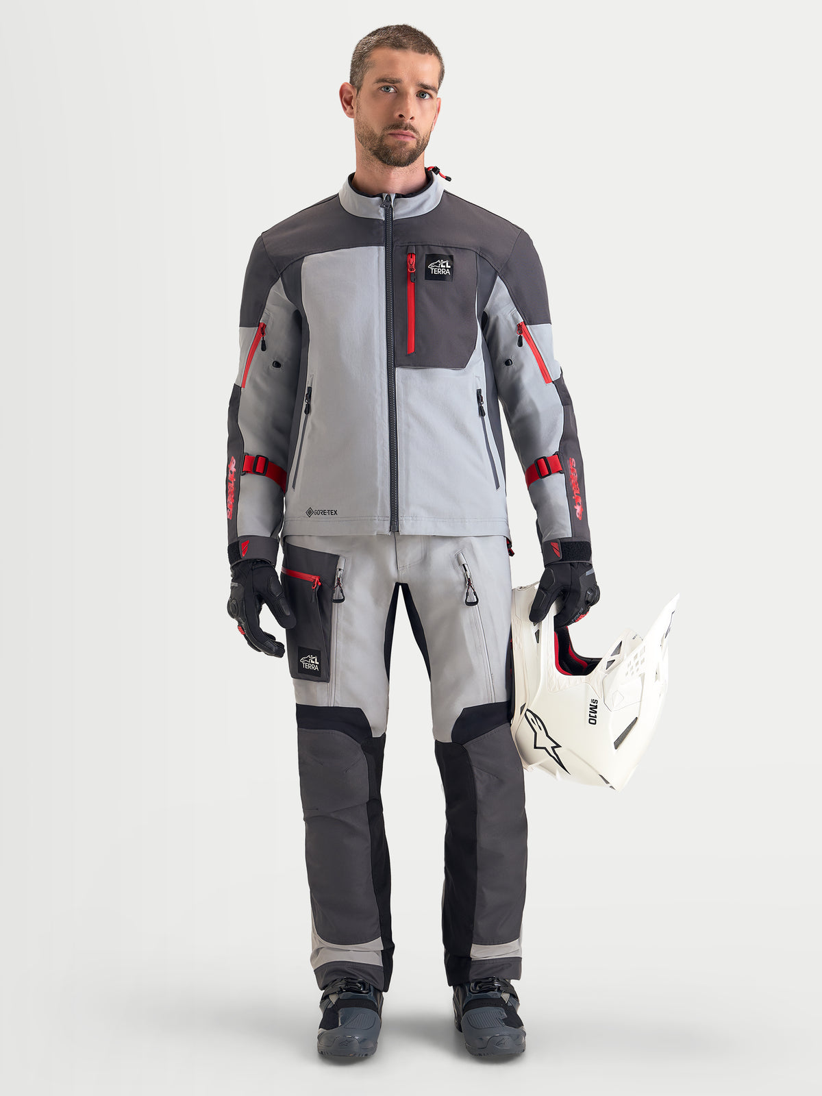 Alpinestars Arenis Gore-Tex uitrustingsset, heren adventure touring motorpak in grijs en donkergrijs met rode accenten, vooraanzicht van model in bijpassende motorkleding en handschoenen die een witte helm vasthoudt