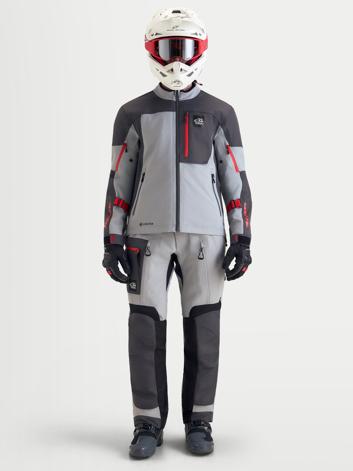 Alpinestars Arenis Gore-Tex uitrustingsset, adventure motorkleding voor heren bestaande uit een motorjas en motorbroek in grijs, donkergrijs en rood, vooraanzicht op een model met bijbehorende kleding en helm, waterdichte all-terrain rijuitrusting
