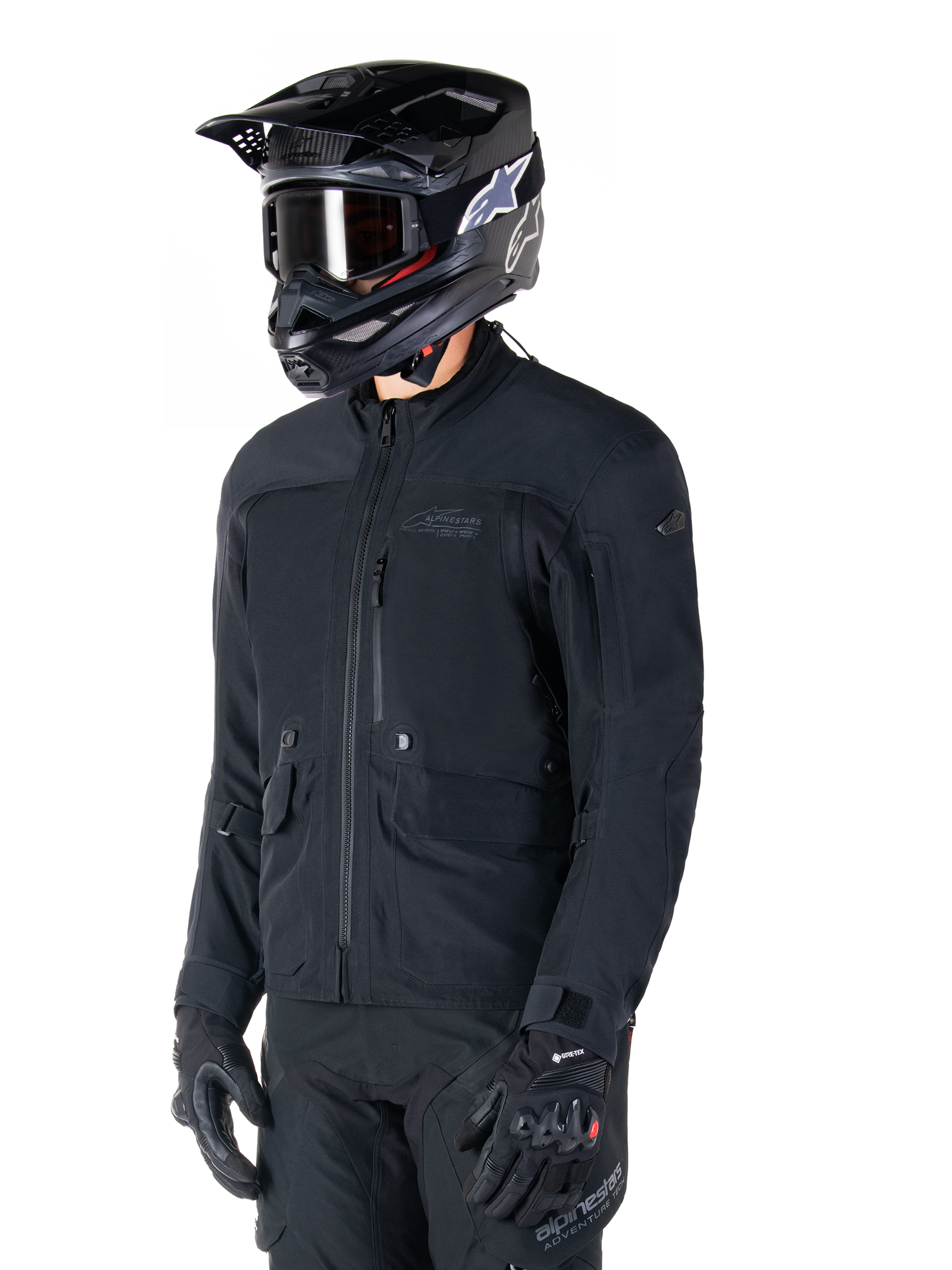 Alpinestars St-7 Prime GORE-TEX jas, zwarte motorjas, gedragen door een rijder met zwarte helm en motorbril, voorzien van grote cargozakken op de borst en een gelamineerde waterdichte buitenlaag, premium uitrusting voor adventure touring en woon-werkverkeer