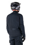 Alpinestars St-7 Prime GORE-TEX Jas, zwart, achteraanzicht van een rijder die de jas draagt met een bijpassende helm en broek, voorzien van Alpinestars-branding en geografische coördinaten op de onderrug.