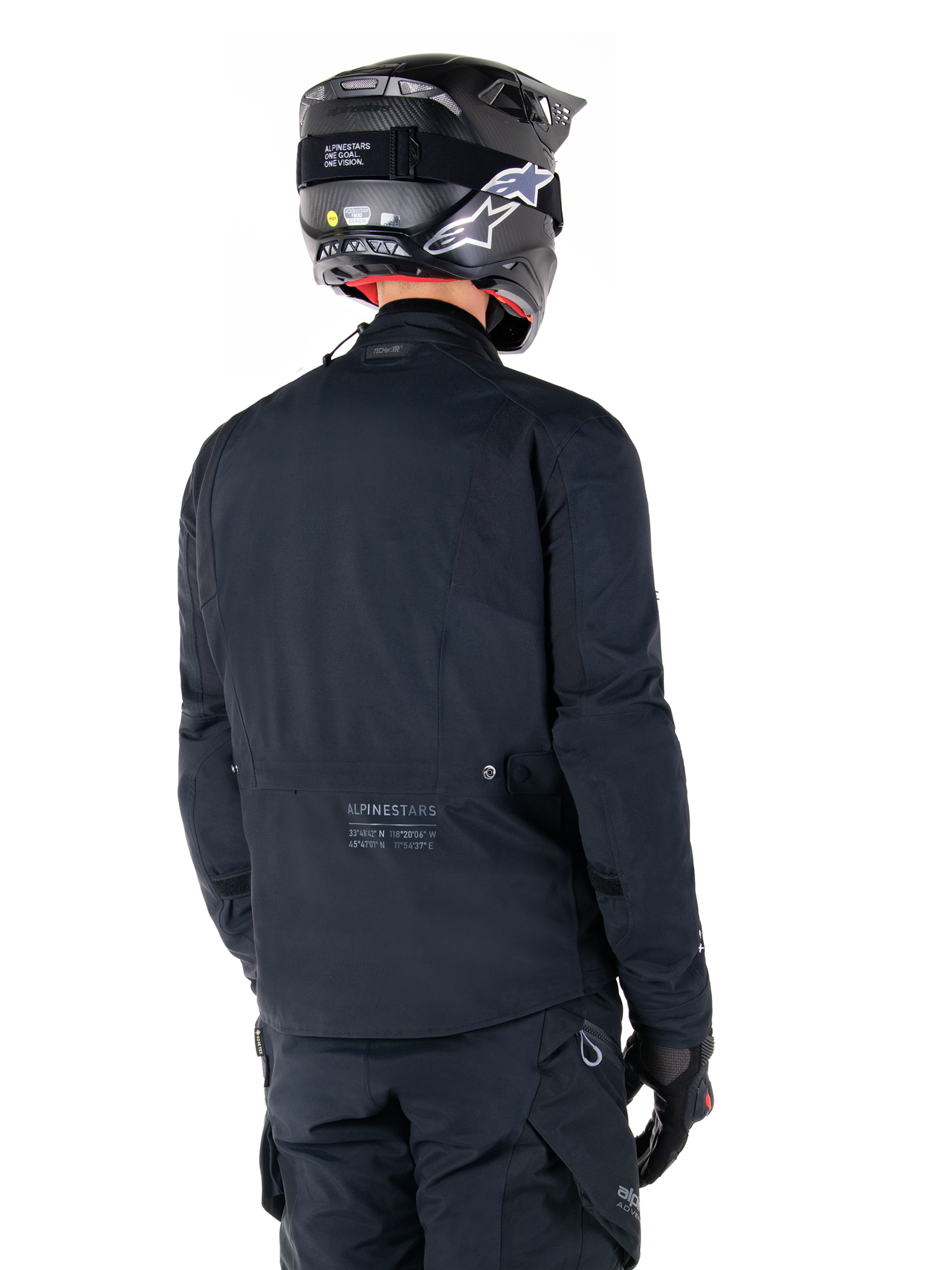 Alpinestars St-7 Prime GORE-TEX Jas, zwart, achteraanzicht van een rijder die de jas draagt met een bijpassende helm en broek, voorzien van Alpinestars-branding en geografische coördinaten op de onderrug.