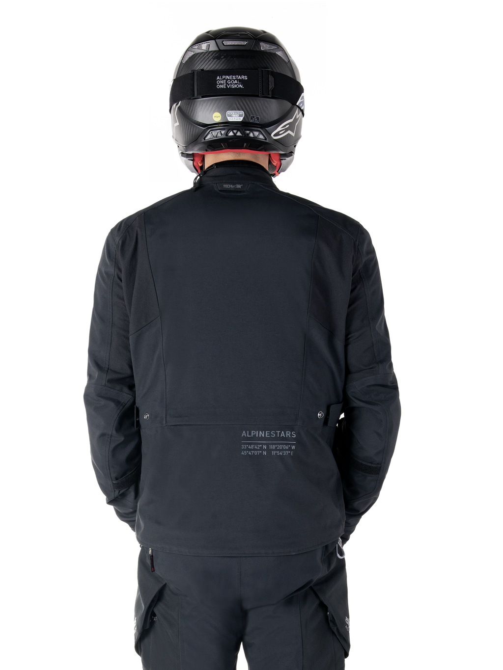 Alpinestars St-7 Prime GORE-TEX Jas, zwart, achteraanzicht van een motorrijder, met subtiel logo en geografische coördinaten op de onderrug, gedragen met een bijpassende zwarte helm en broek, strakke en technische motorkleding