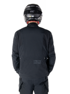 Alpinestars St-7 Prime GORE-TEX Jas, zwart, achteraanzicht van een motorrijder, met subtiel logo en geografische coördinaten op de onderrug, gedragen met een bijpassende zwarte helm en broek, strakke en technische motorkleding