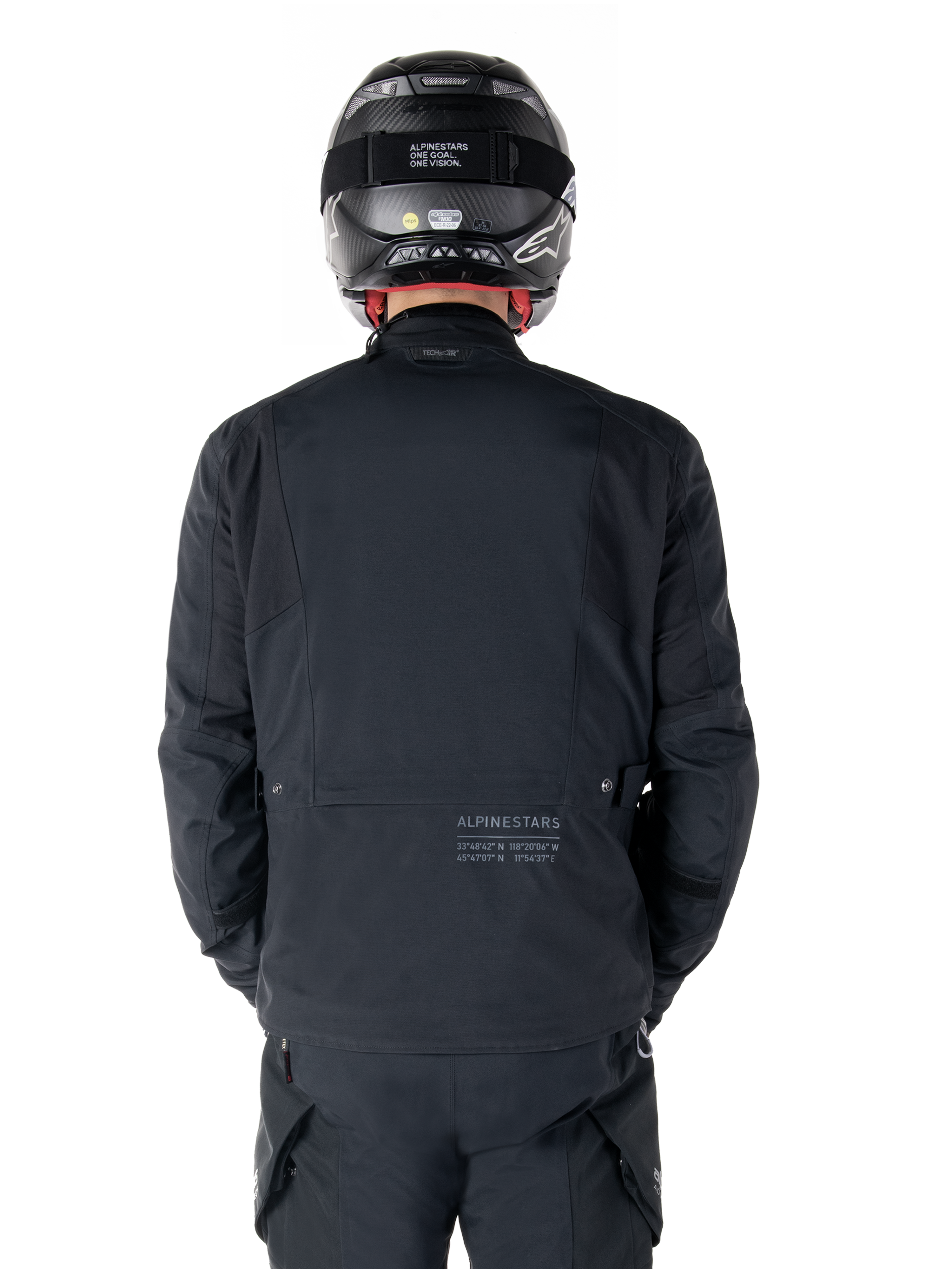 Alpinestars St-7 Prime GORE-TEX Jas, zwart, achteraanzicht van een motorrijder, met subtiel logo en geografische coördinaten op de onderrug, gedragen met een bijpassende zwarte helm en broek, strakke en technische motorkleding