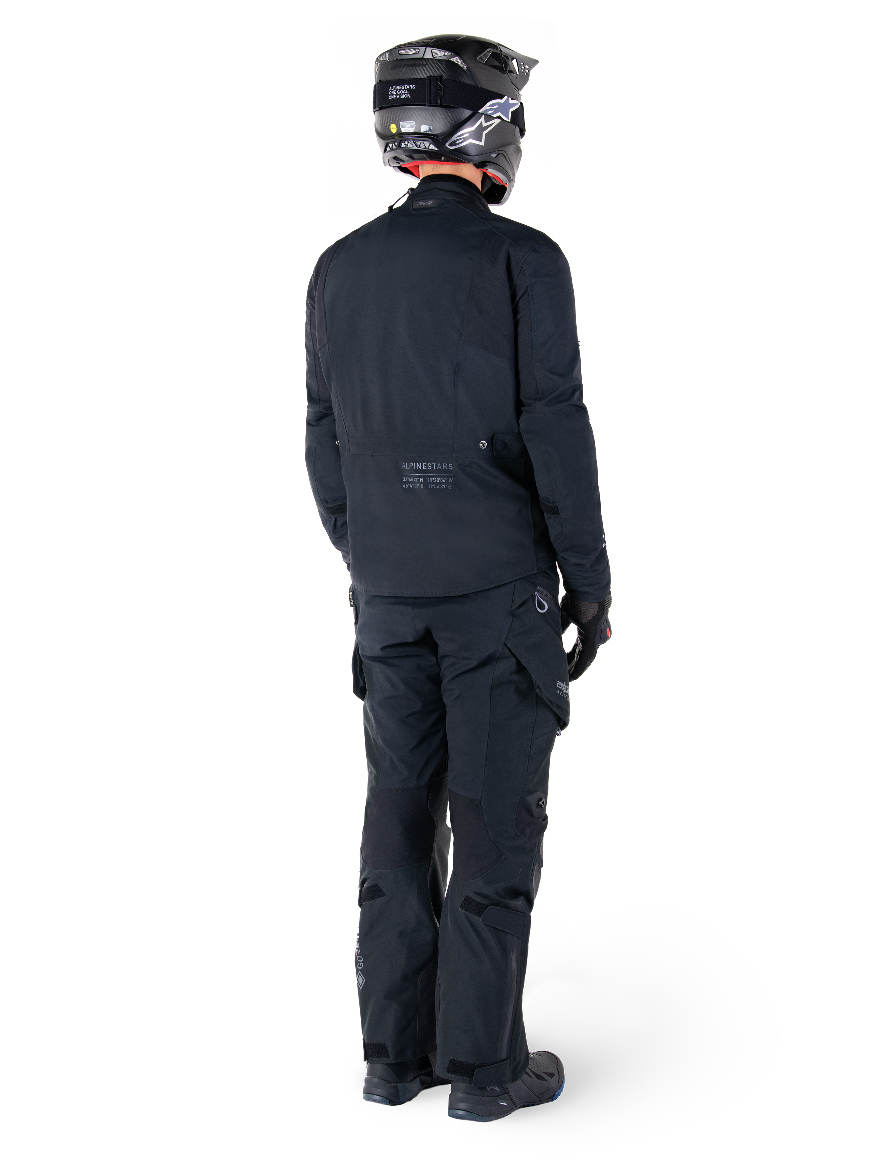 Achteraanzicht van een motorrijder die de Alpinestars St-7 Prime GORE-TEX Jas in het zwart draagt, gecombineerd met een bijpassende zwarte toerbroek, een zwarte motorhelm met wit logo en zwarte motorlaarzen, volledige beschermende adventure-uitrusting voor toeren en woon-werkverkeer.