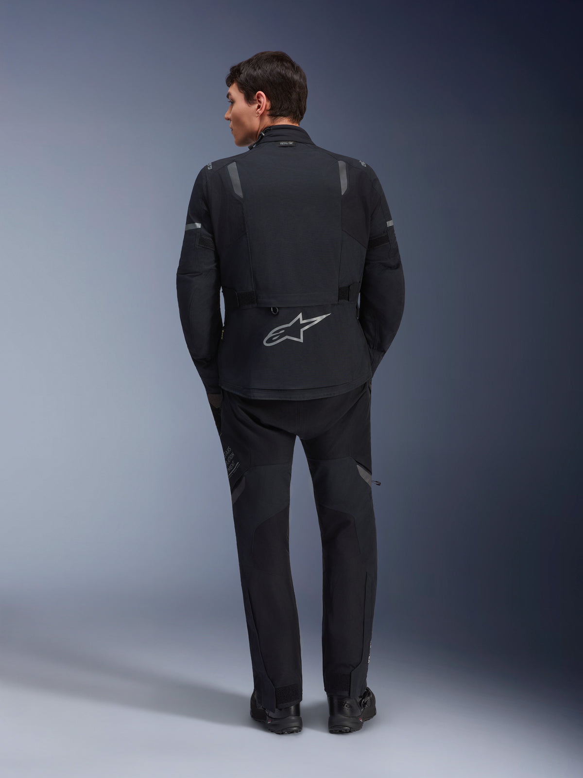 Alpinestars ST-7 2L GORE-TEX jas en bijbehorende ST-7 2L GORE-TEX broek, zwarte en donkergrijze adventure motoruitrusting, achteraanzicht met reflecterend Alpinestars-logo op de jas en technische textielconstructie