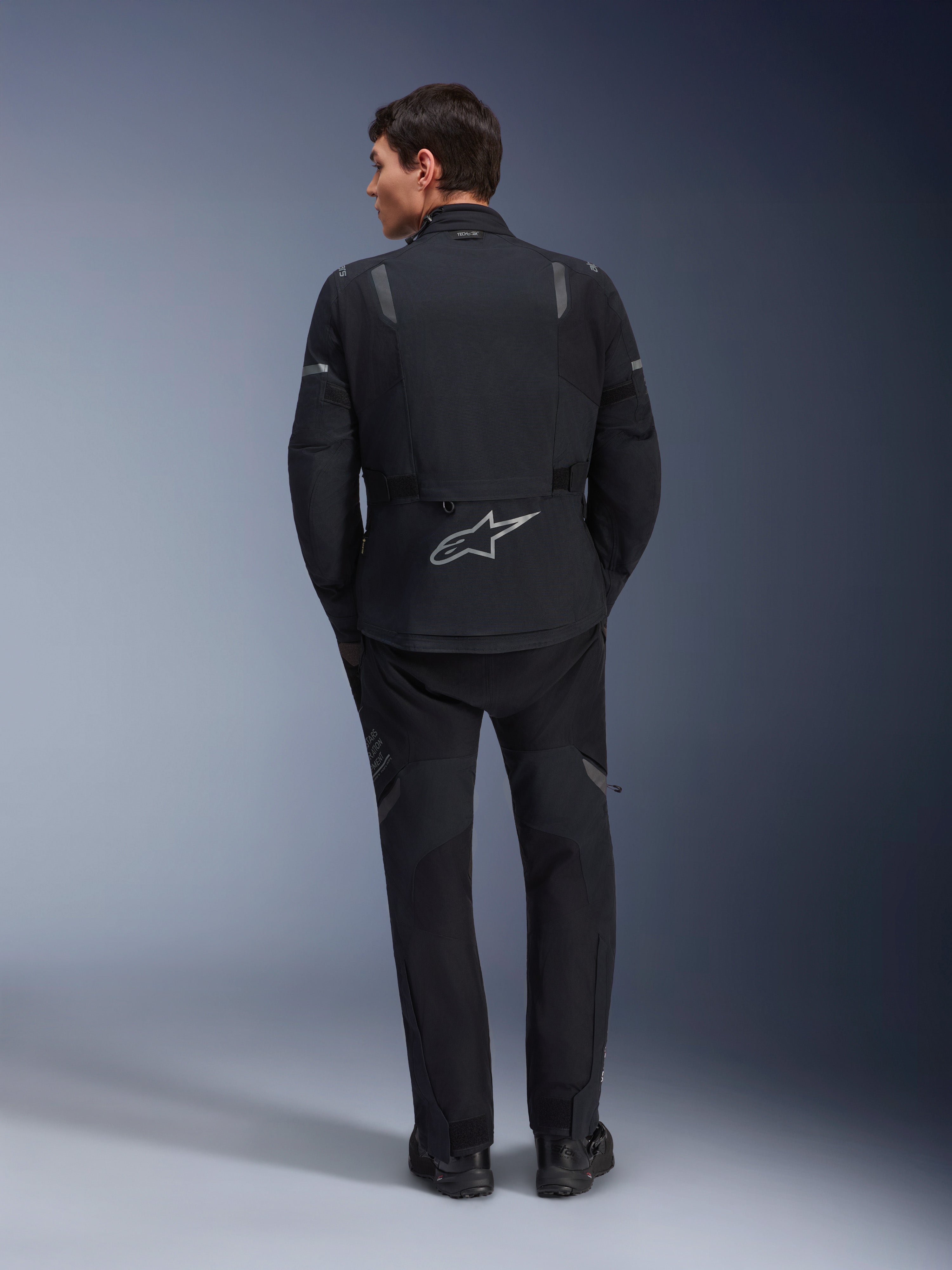Alpinestars ST-7 2L GORE-TEX Jas, adventure motorjas, zwart en donkergrijs, achteraanzicht van een man die de jas en een bijpassende zwarte broek draagt, voorzien van een groot reflecterend Astar-logo op de onderrug, waterdichte textieluitrusting voor touring en rijden op de weg
