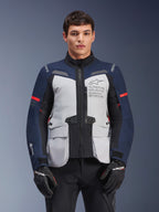 Alpinestars ST-7 2L GORE-TEX jas, motor adventure touringjas, ijsgrijs en donkerblauw met rode accenten, waterdicht en ademend, gedragen door een model met zwarte handschoenen, voorzien van Alpinestars Exploration Equipment-branding op de borst
