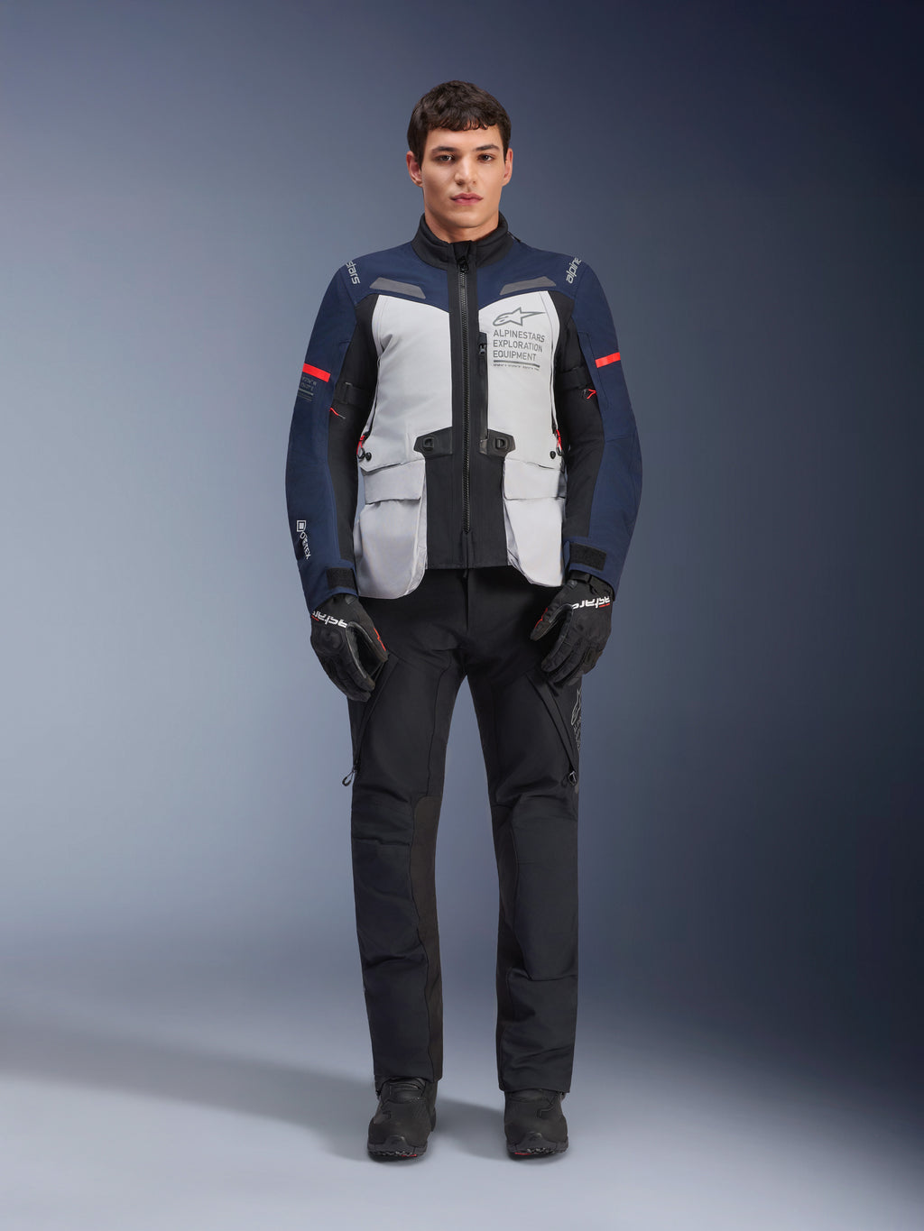 Alpinestars ST-7 2L GORE-TEX jas, adventure touring motorjas, Ice Gray, donkerblauw en zwart, gedragen met een zwarte broek en handschoenen, voorzien van grote cargozakken aan de voorzijde, rode accenten op de mouwen en een gelamineerde GORE-TEX buitenlaag voor waterdichte prestaties.