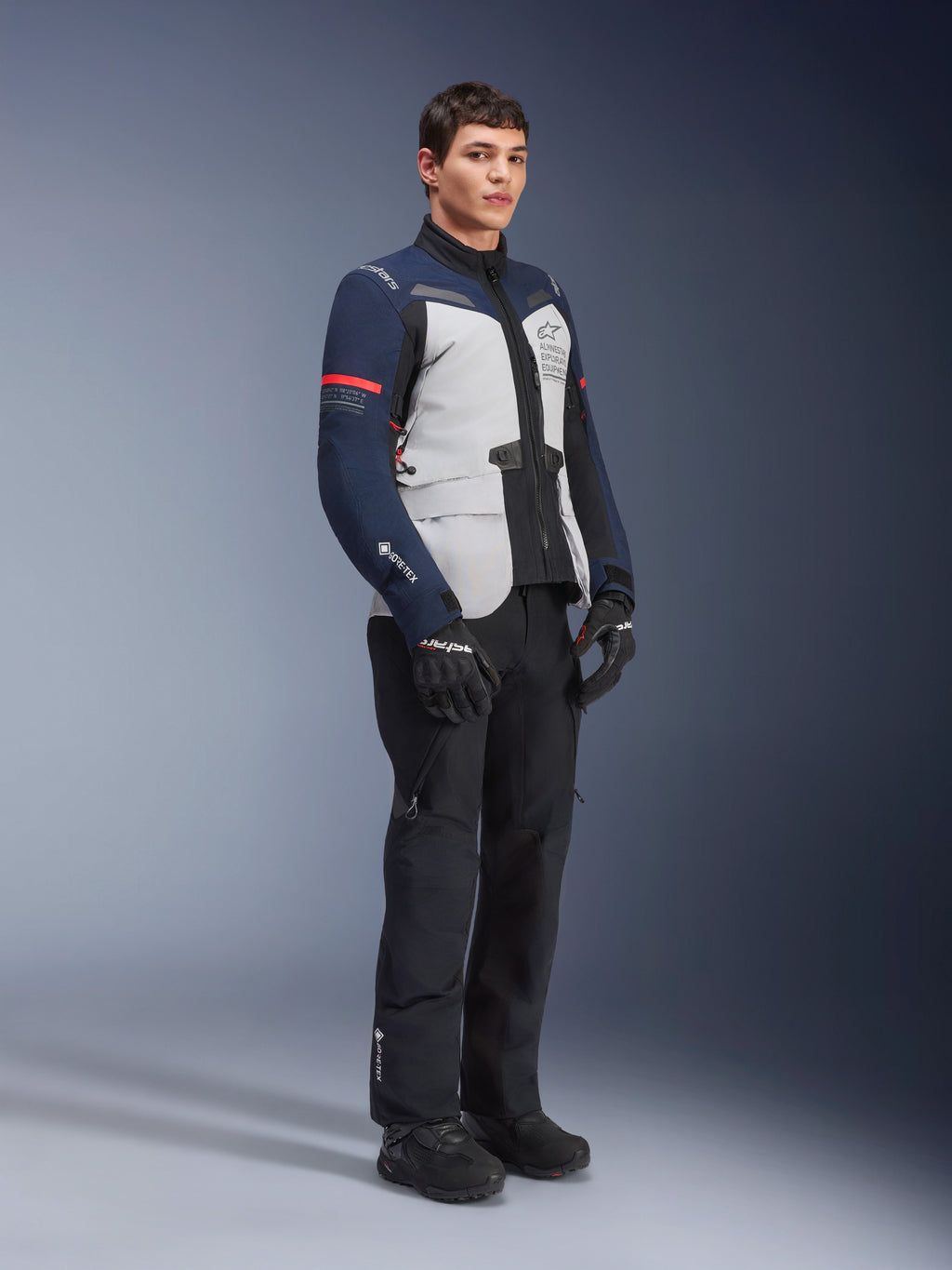Alpinestars ST-7 2L GORE-TEX jas in de kleuren Ice Gray, donkerblauw en zwart met rode accenten, gedragen door een mannelijk model met zwarte motorbroek en handschoenen, professionele adventure touring motoruitrusting