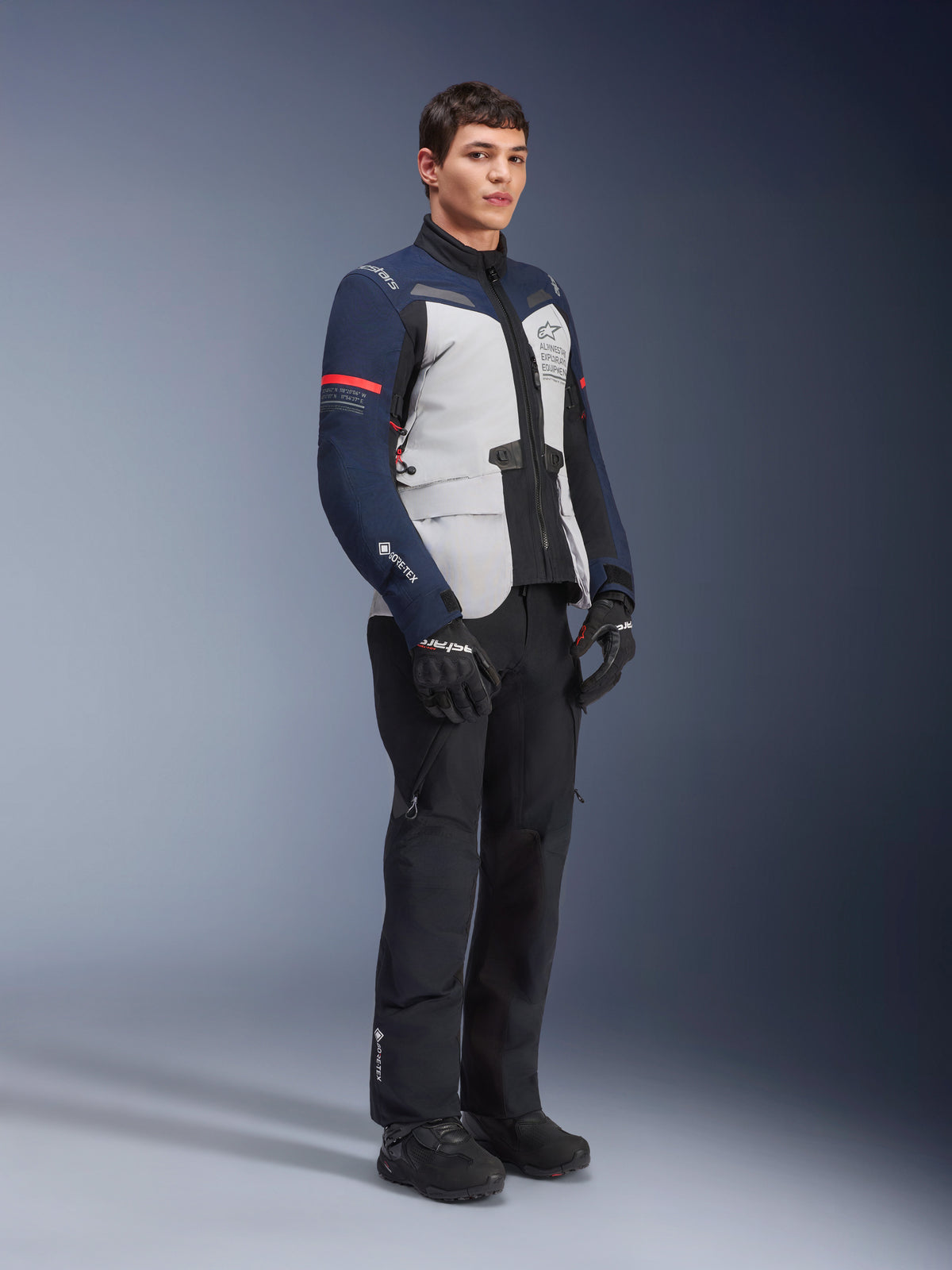 Alpinestars ST-7 2L GORE-TEX jas in de kleuren Ice Gray, donkerblauw en zwart met rode accenten, gedragen door een mannelijk model met zwarte motorbroek en handschoenen, professionele adventure touring motoruitrusting