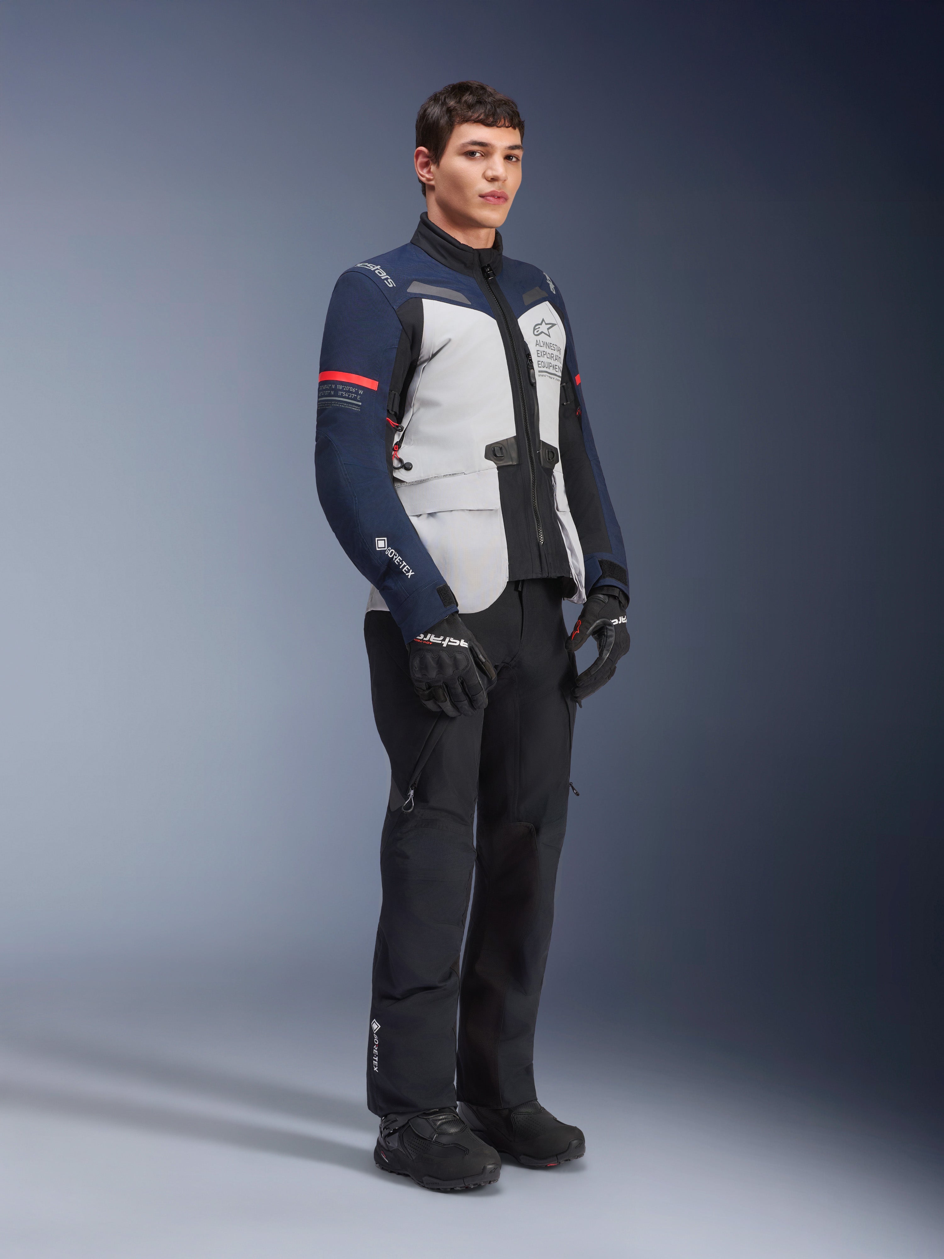 Alpinestars ST-7 2L GORE-TEX jas in de kleuren Ice Gray, donkerblauw en zwart met rode accenten, gedragen door een mannelijk model met zwarte motorbroek en handschoenen, professionele adventure touring motoruitrusting
