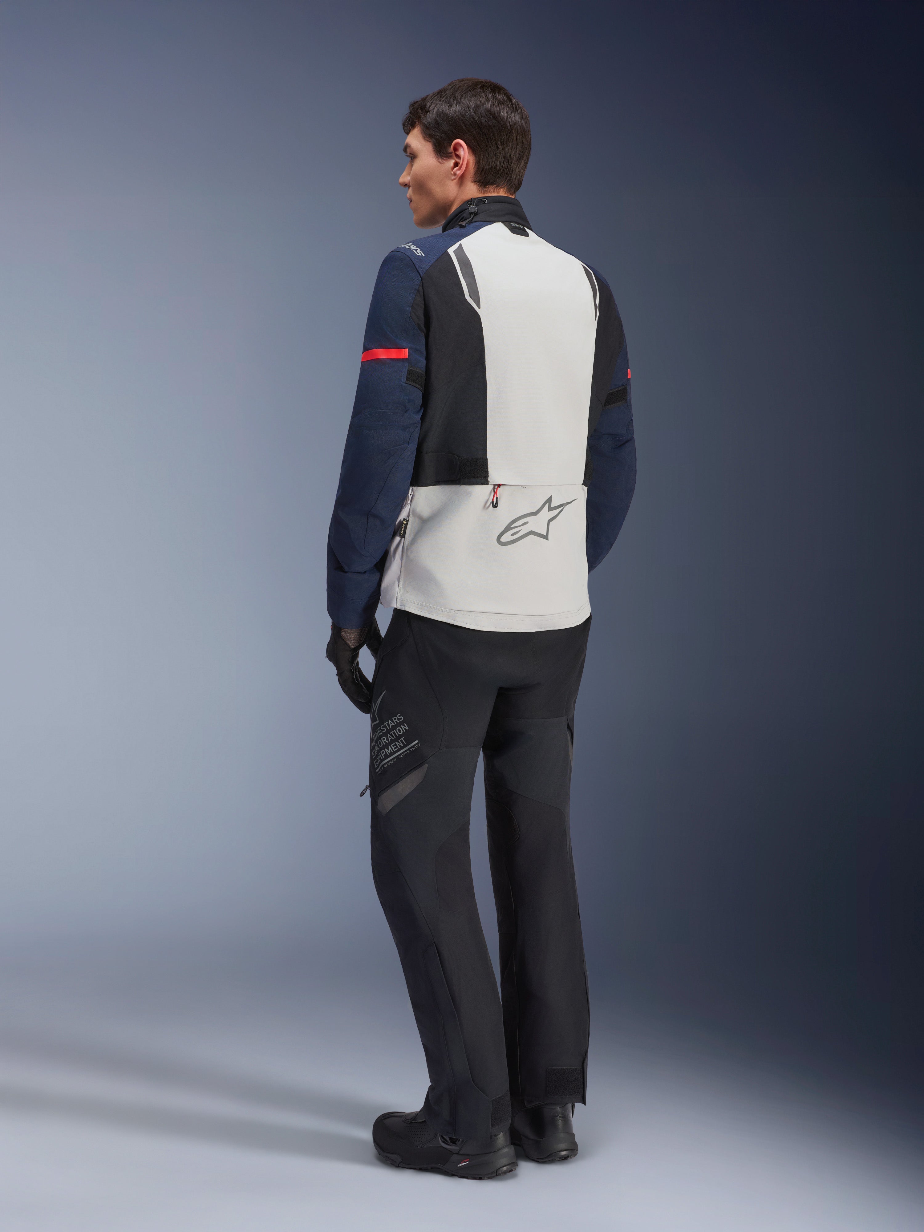 Model draagt Alpinestars ST-7 2L GORE-TEX Jas in Ice Gray, donkerblauw en zwart, achteraanzicht, lichtgrijs rugpaneel met Alpinestars-logo, donkerblauwe mouwen met rode streep, gecombineerd met zwarte adventure motorbroek en laarzen, motor touring uitrusting