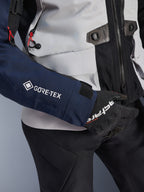 Alpinestars ST-7 2L GORE-TEX Jas, close-up van donkerblauwe mouw met wit GORE-TEX logo, ijsgrijs torso met zwarte details en rode koordaccenten, gedragen met zwarte Alpinestars handschoenen en broek, adventure touring motorkleding