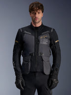Alpinestars ST-7 2L GORE-TEX jas, heren adventure touring motorjas, donkergrijs en zwart met khaki accenten, voorzien van 'Alpinestars Exploration Equipment' branding, waterdichte cargozakken en een hoge kraag, gedragen door een model met zwarte handschoenen