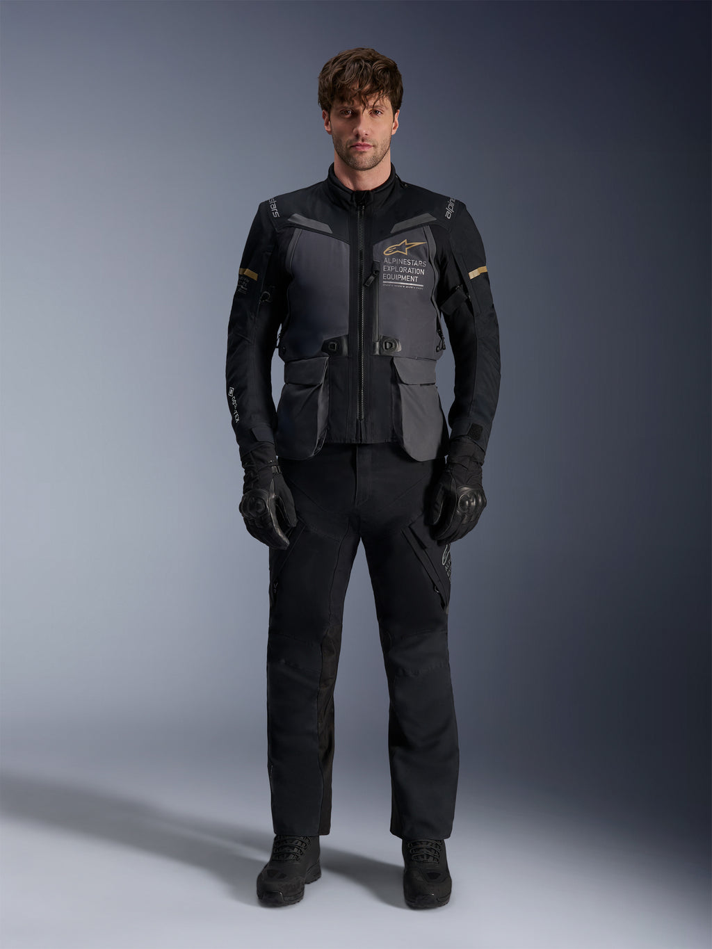 Mannelijk model draagt de Alpinestars ST-7 2L GORE-TEX Jas in Deep Gray Black Dark Khaki, voorzien van 'ALPINESTARS EXPLORATION EQUIPMENT' branding op de borst, gecombineerd met bijpassende zwarte motorbroek, handschoenen en laarzen, studio vooraanzicht.
