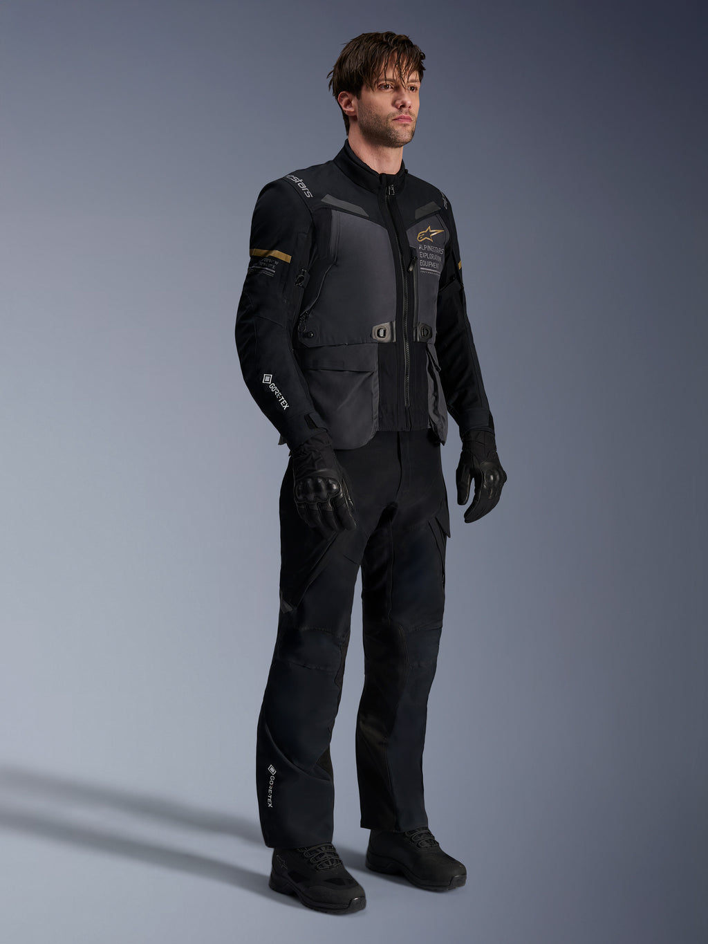 Mannelijk model in de Alpinestars ST-7 2L GORE-TEX Jas in Deep Gray Black en Dark Khaki, gecombineerd met bijpassende zwarte adventure-broek en handschoenen, voorzien van een waterdichte gelamineerde buitenlaag met cargozakken en technische branding, ideaal voor adventure touring.