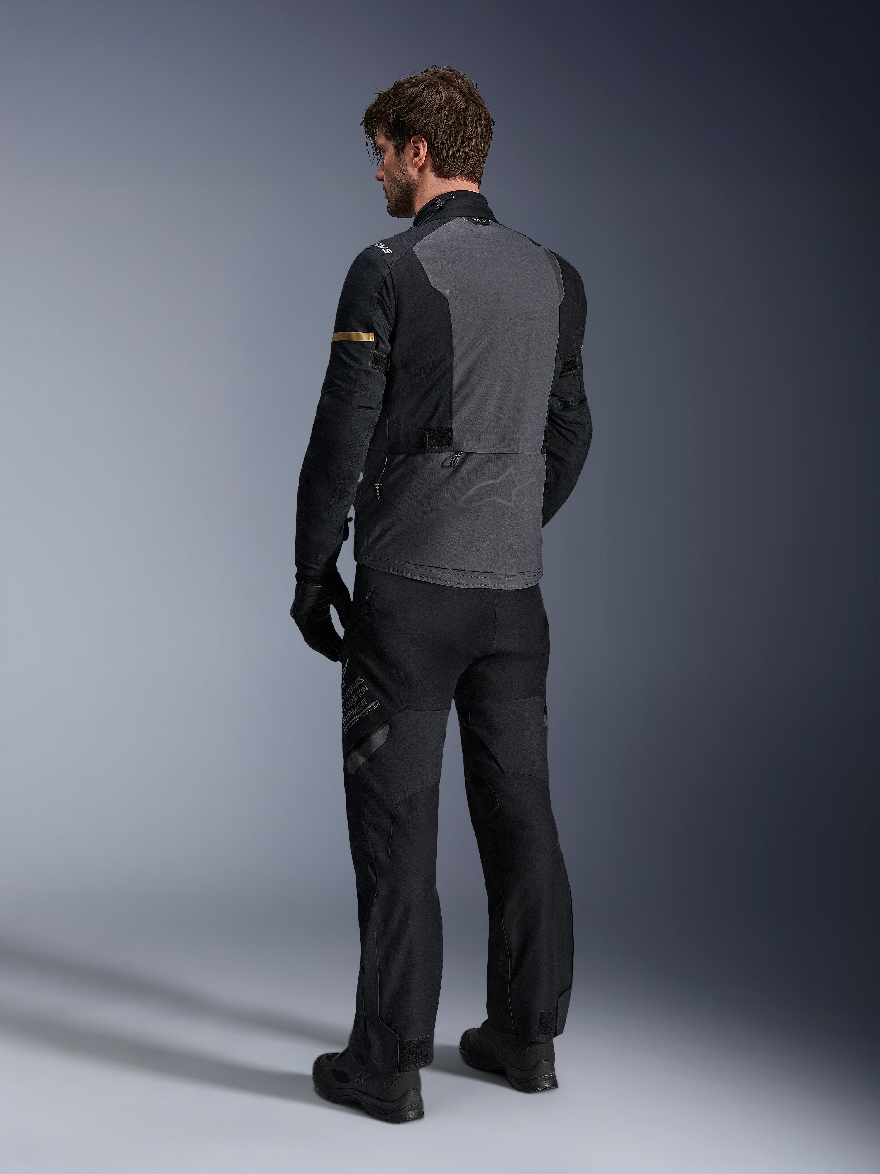 Alpinestars ST-7 2L GORE-TEX Jas, donkergrijs en zwart met Dark Khaki accenten, achteraanzicht van een motorrijder die de jas en bijpassende motorbroek draagt, adventure touring uitrusting met waterdichte gelamineerde constructie