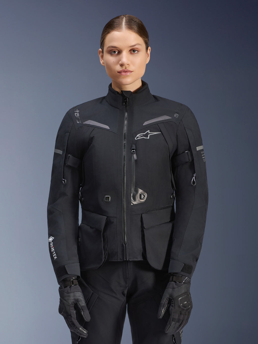 Vrouw draagt Alpinestars Stella ST-7 2L GORE-TEX jas, zwart met donkergrijze accenten, motor touring jas met grote cargozakken en hoge kraag, gecombineerd met zwarte handschoenen en broek, waterdichte adventure motorkleding