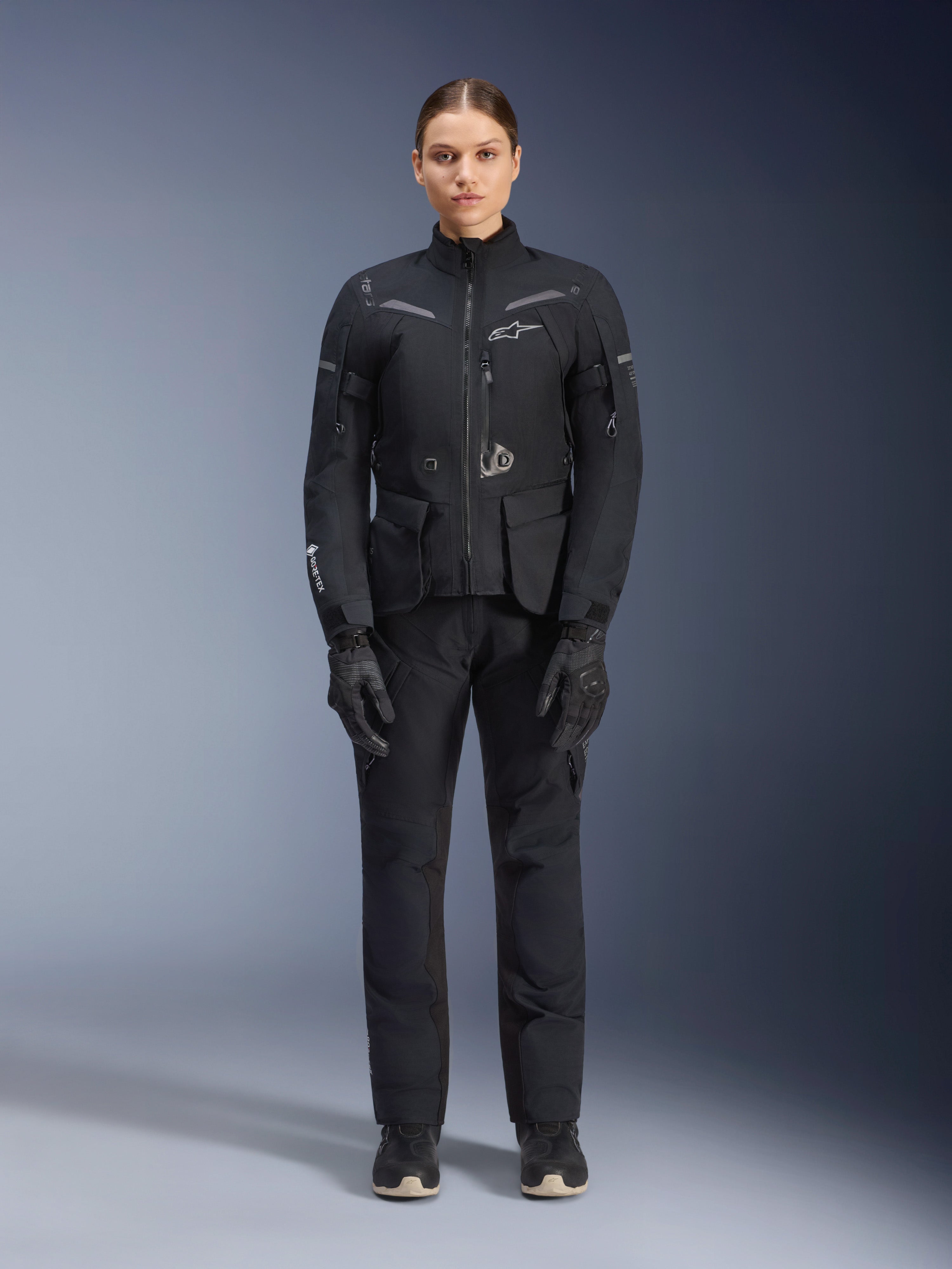 Vrouwelijke motorrijder draagt Alpinestars Stella ST-7 2L GORE-TEX jas en broek voor dames, zwarte touring motoroutfit, waterdichte en ademende technische kleding, gecombineerd met bijpassende zwarte handschoenen en laarzen, volledige beschermende uitrusting met reflecterende elementen, modern technisch ontwerp voor adventure rijden