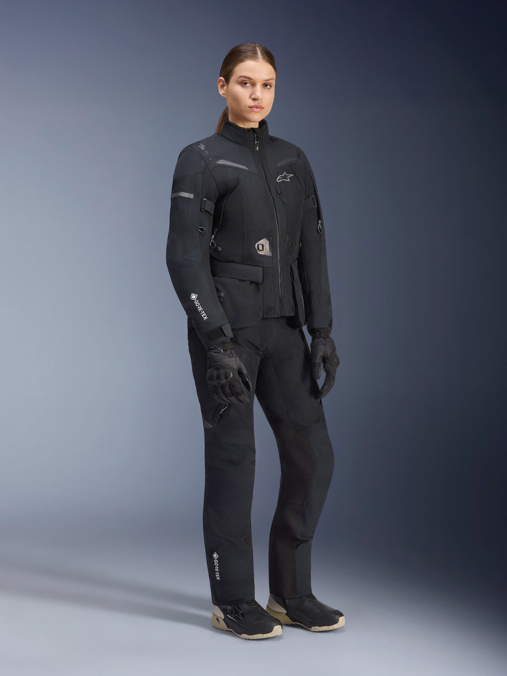 Vrouwelijk model in Alpinestars Stella ST-7 2L GORE-TEX adventure touring-pak, zwart en donkergrijs jack en broek, met opvallende GORE-TEX-branding op de mouw en broekspijp, gecombineerd met zwarte handschoenen en motorlaarzen, professionele motoruitrusting voor touring onder alle weersomstandigheden