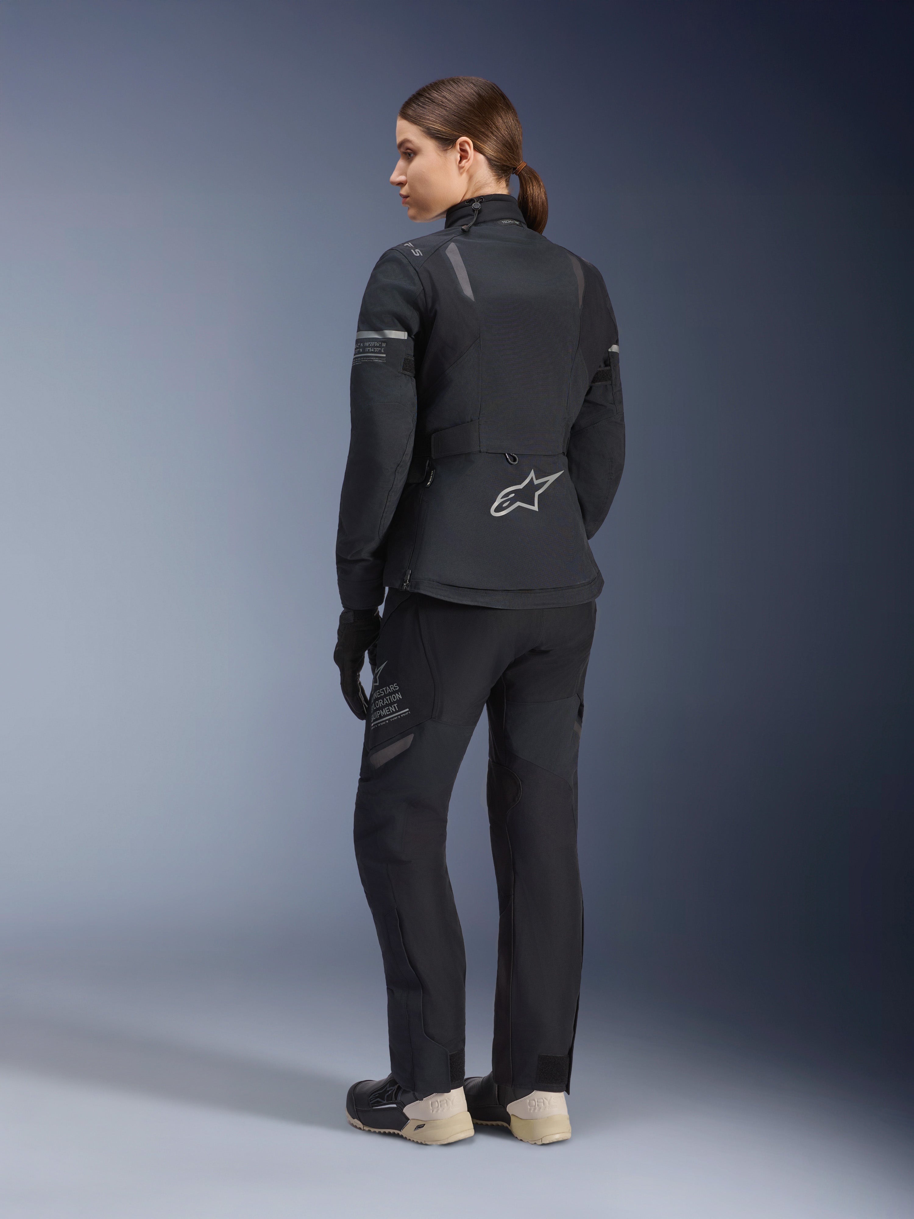 Alpinestars Stella ST-7 2L GORE-TEX damesjas, zwart en donkergrijs, adventure touring jas van achteren gezien, gedragen met bijpassende zwarte broek en crème laarzen, voorzien van het Alpinestars-logo op de onderrug, ontworpen voor toerrijden in alle seizoenen